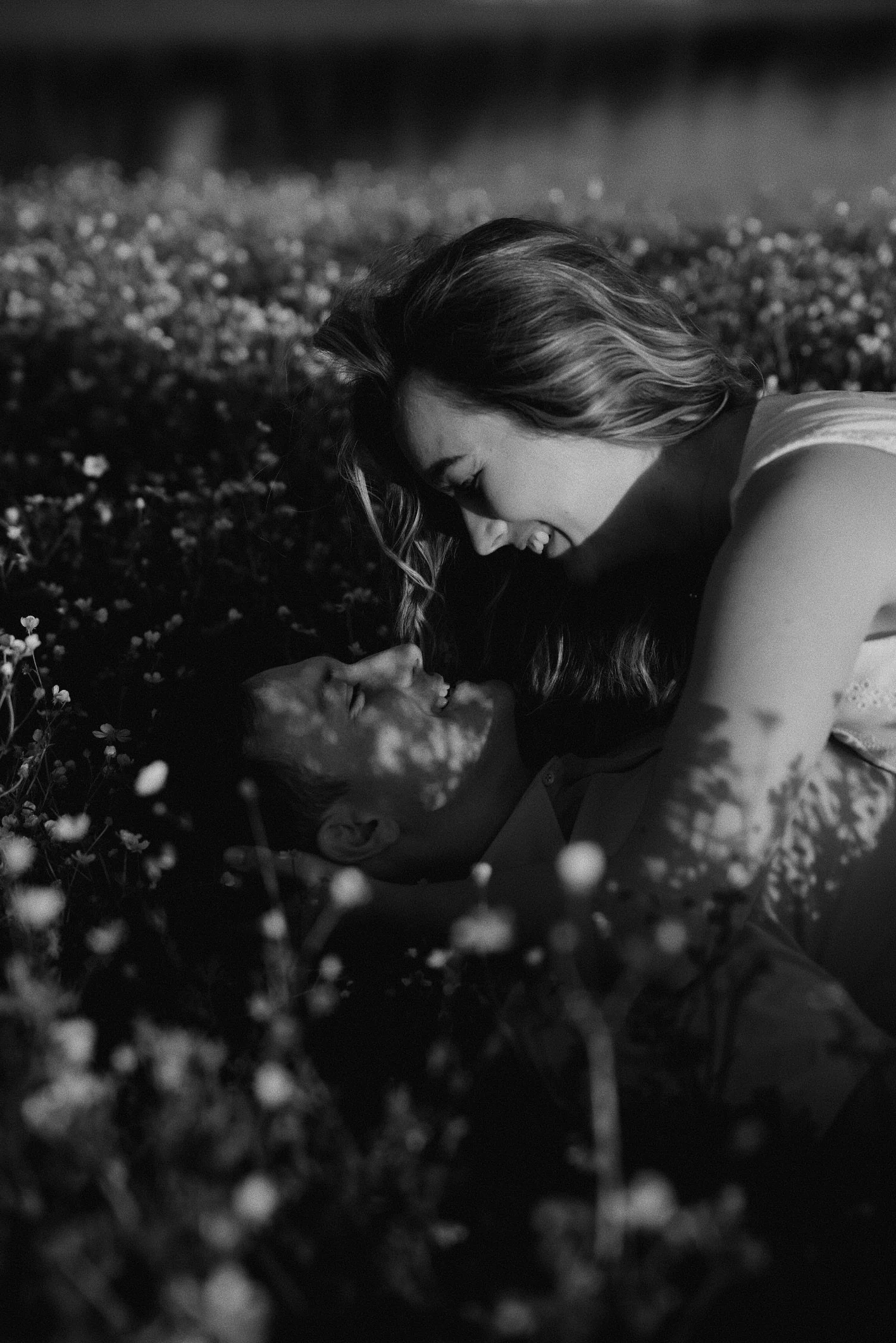 A black and white photo of a woman and a man lying in a field of flowers, smiling and looking at each other, with the woman on top of the man.