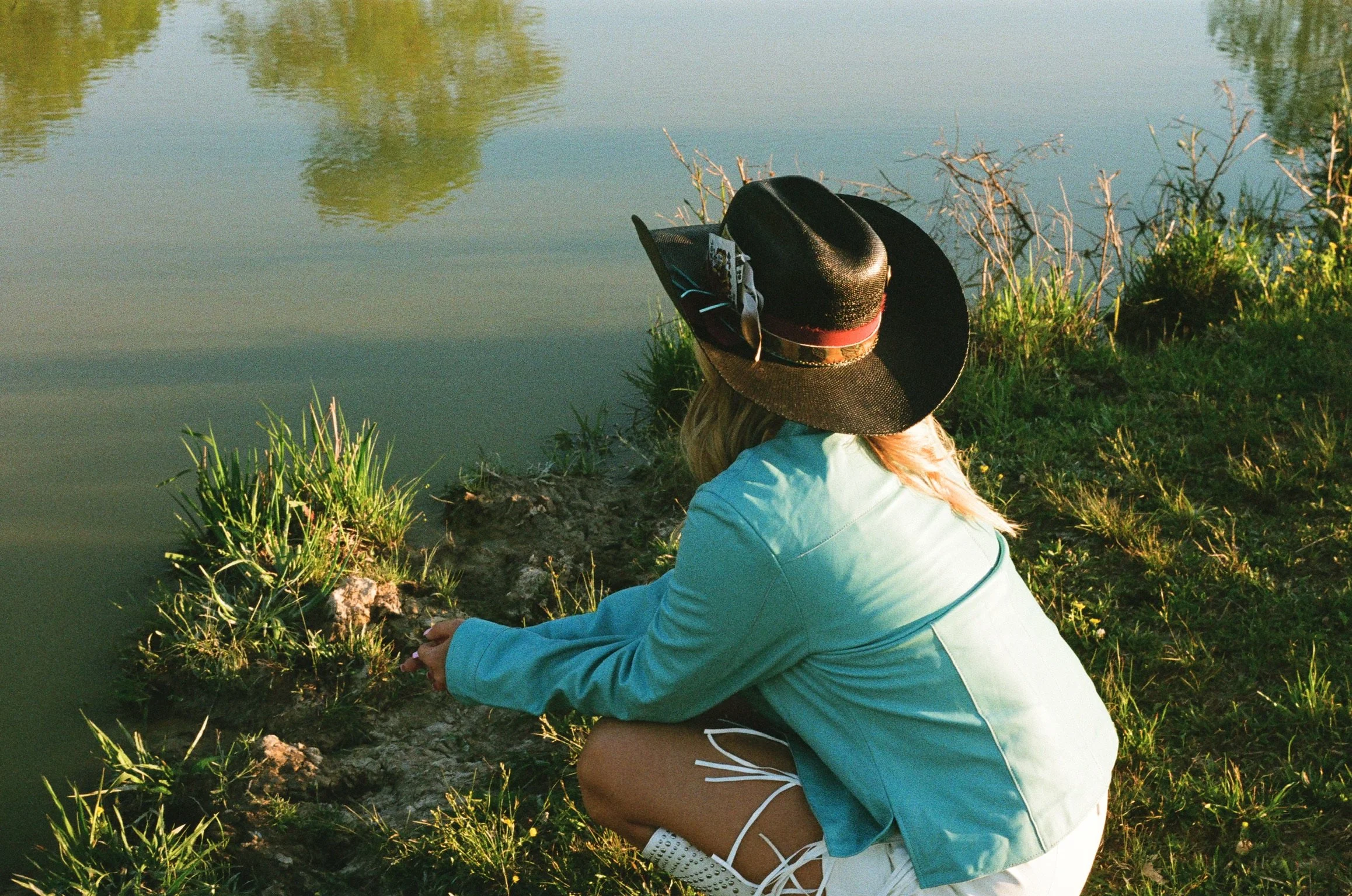 A person wearing a wide-brimmed hat, a light blue jacket, and white sneakers sits by a calm lake or pond, surrounded by green grass and plants, gazing at the water with reflections of trees visible on its surface.