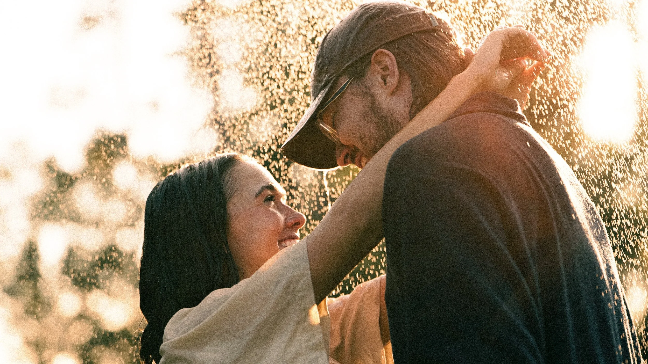 A couple is embracing in the rain with sunlight shining through the water droplets, creating a warm and romantic atmosphere.