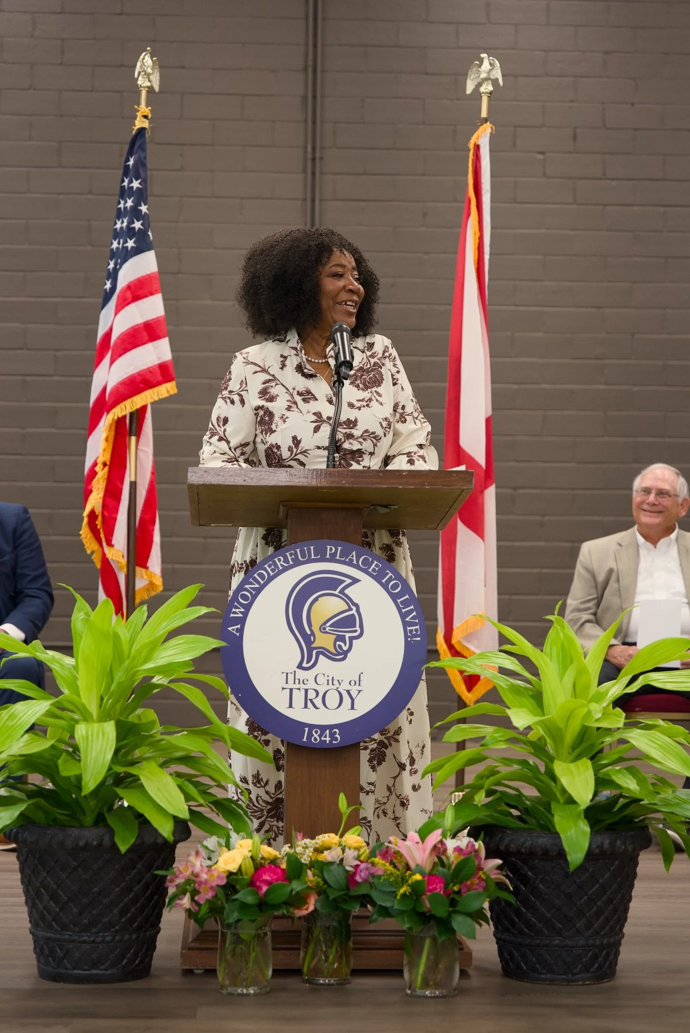 A woman with curly hair speaking at a podium with a city seal for Troy, Michigan. Two American flags flank her and a floral arrangement is in front of the podium. Several people are seated behind her.