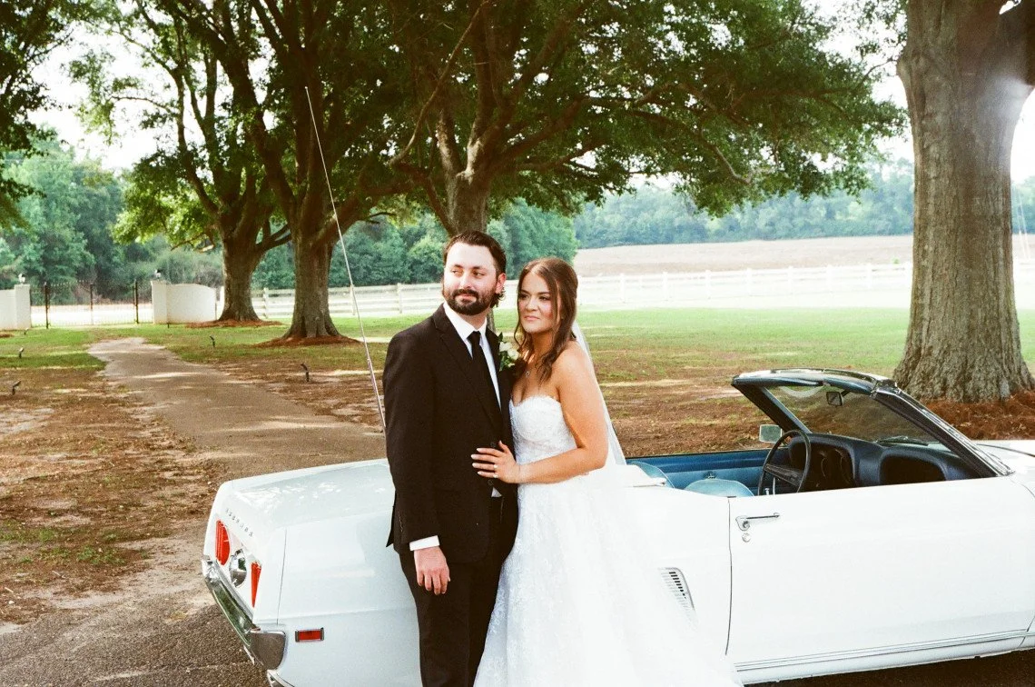 A couple in wedding attire standing next to a white convertible car outdoors with trees and a field in the background.
