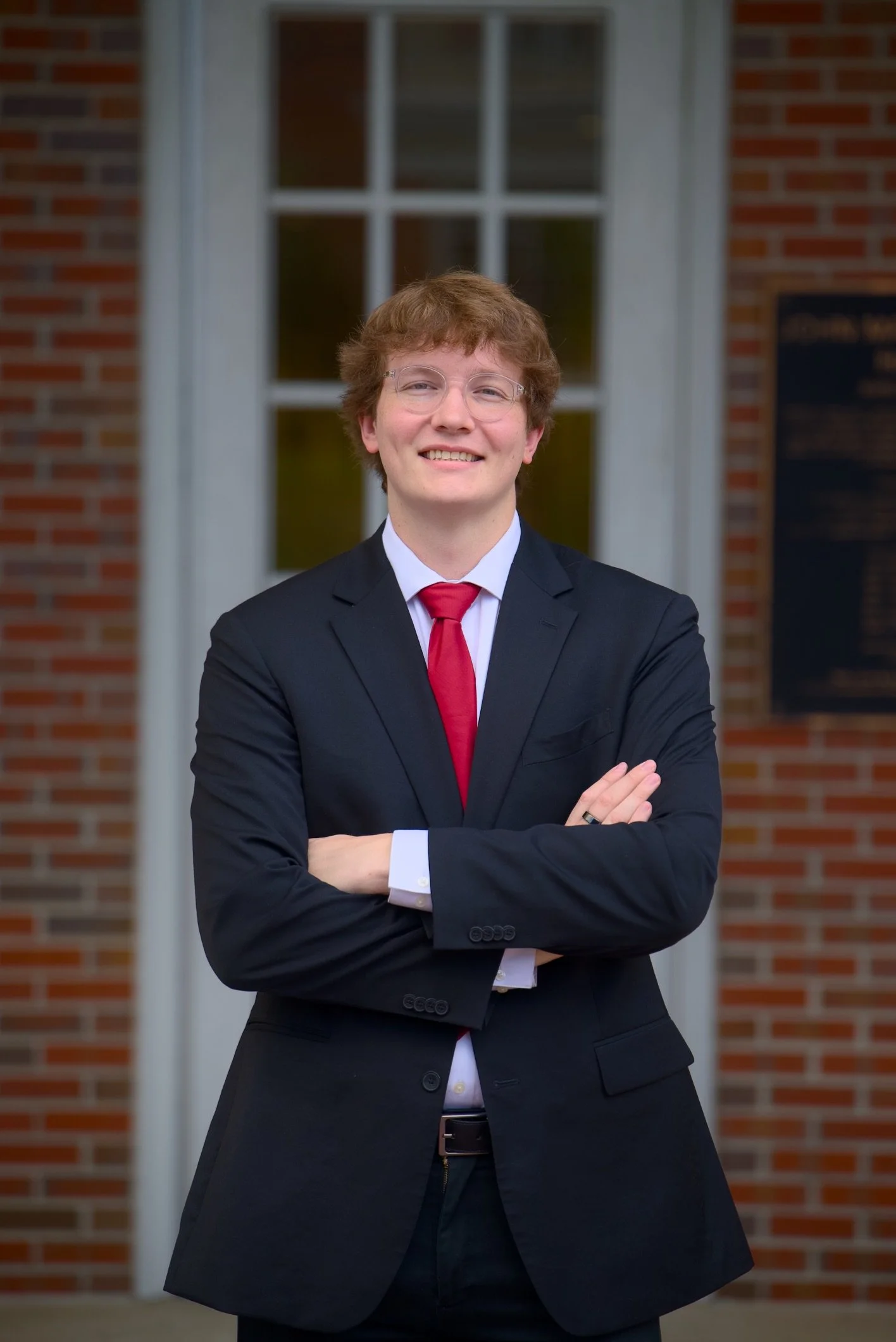 A young man wearing a black suit with a white shirt and red tie, standing outdoors in front of a brick building with a large window, arms crossed, smiling confidently.