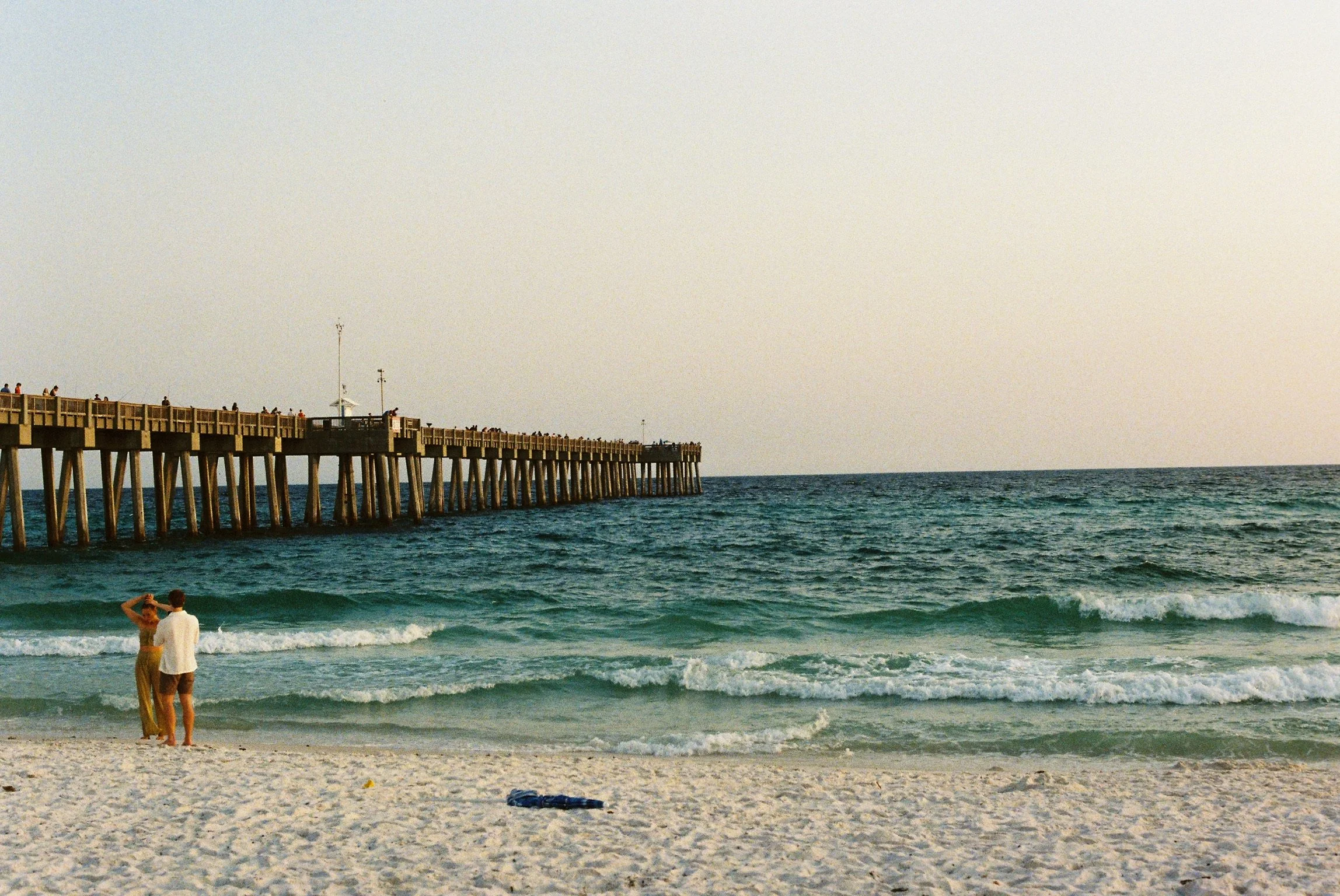 People standing and talking on the beach near the shoreline with a pier extending into the ocean in the background.