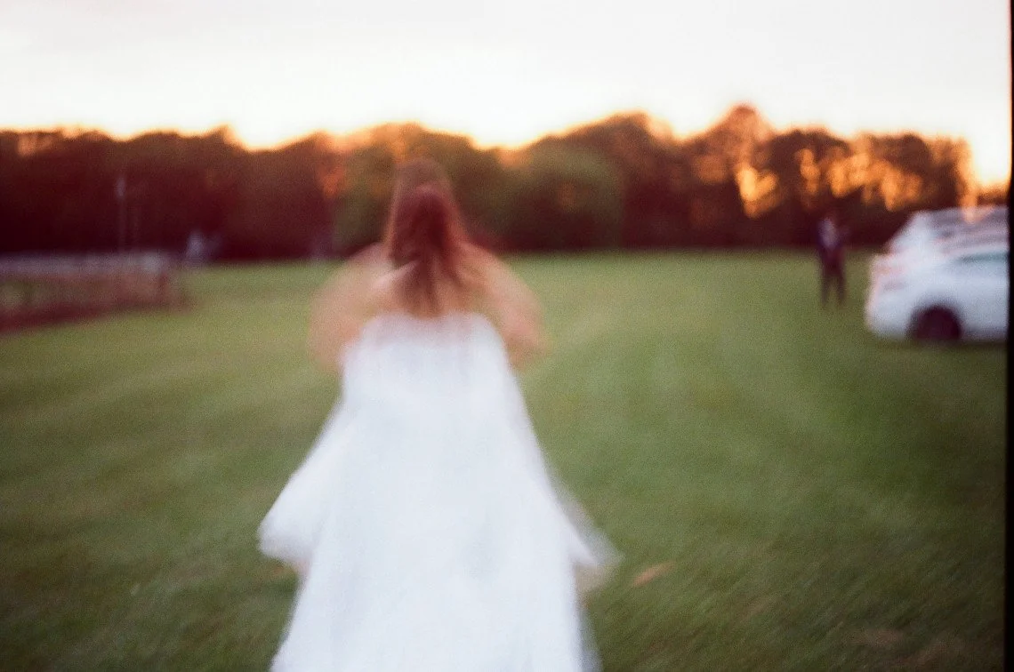 A woman in a white dress walking across a grassy field towards trees at sunset, with a person and a white vehicle in the distance.