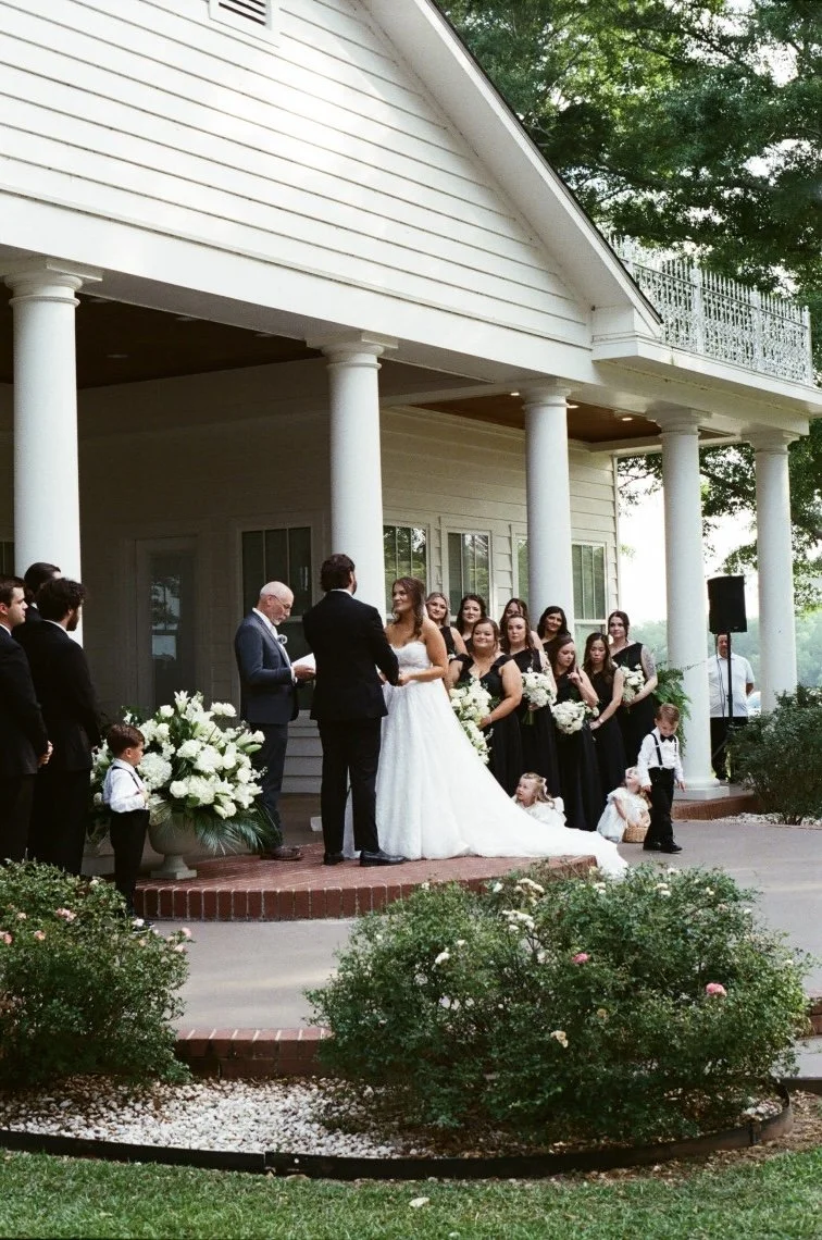 A wedding ceremony taking place outdoors in front of a white house with columns. The bride and groom are at the center, with the bride wearing a white gown and the groom in a black suit. Bridesmaids in black dresses and groomsmen in black suits stand