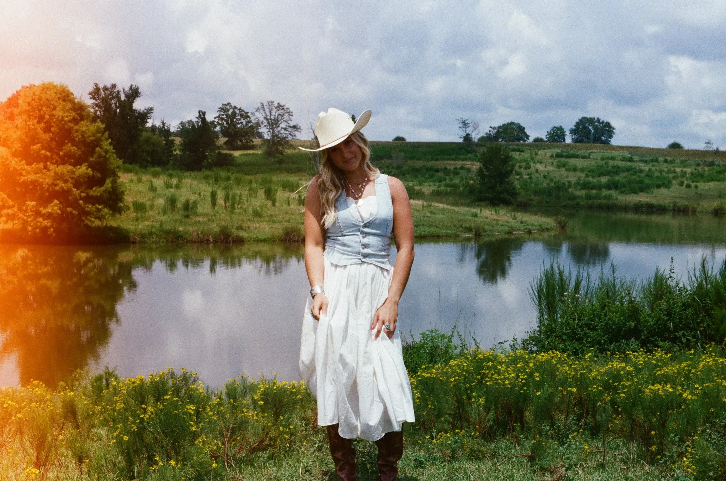 A woman wearing a white cowboy hat, a white top, and a white skirt stands near a river with greenery and trees in the background, under a cloudy sky.