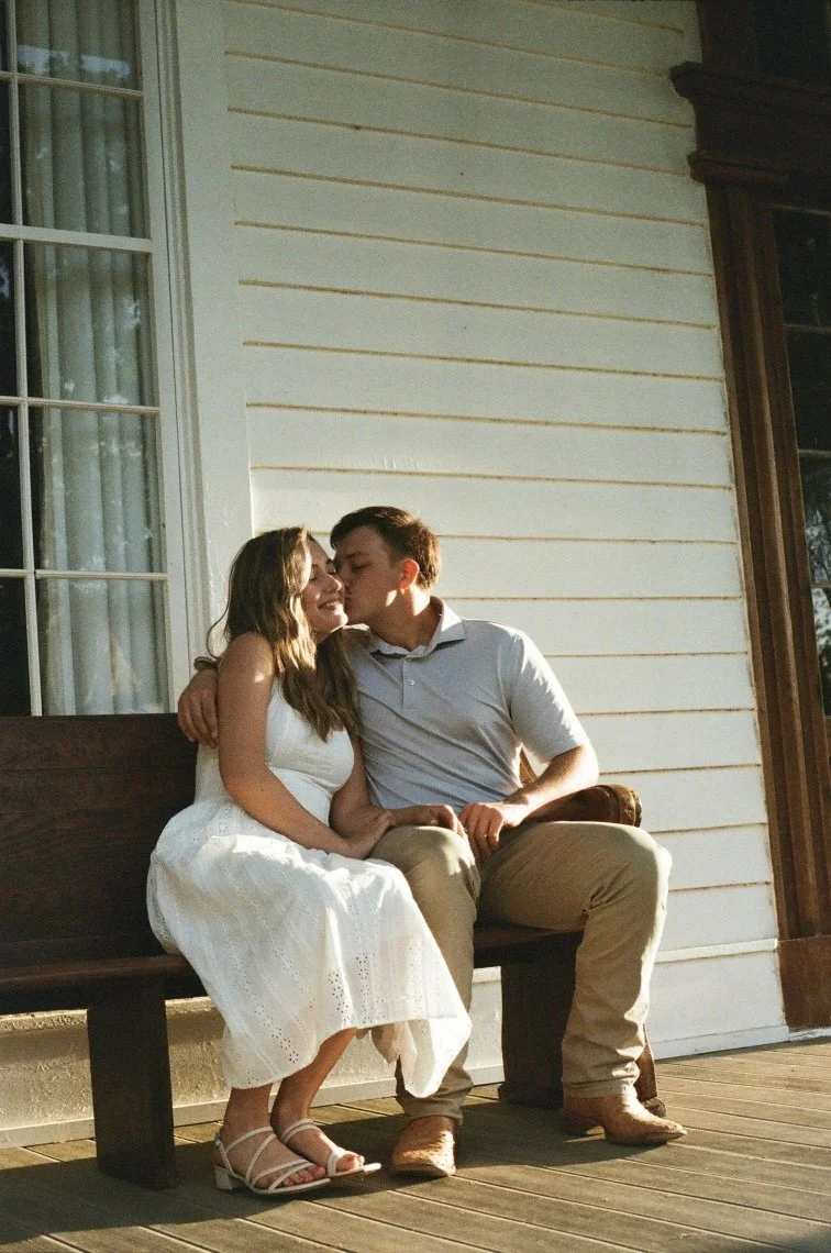A young couple sitting on a bench outside a house. The girl is wearing a white dress and sandals, and the boy is in a light-colored polo shirt and khakis. The boy is kissing the girl's forehead, and they are smiling.