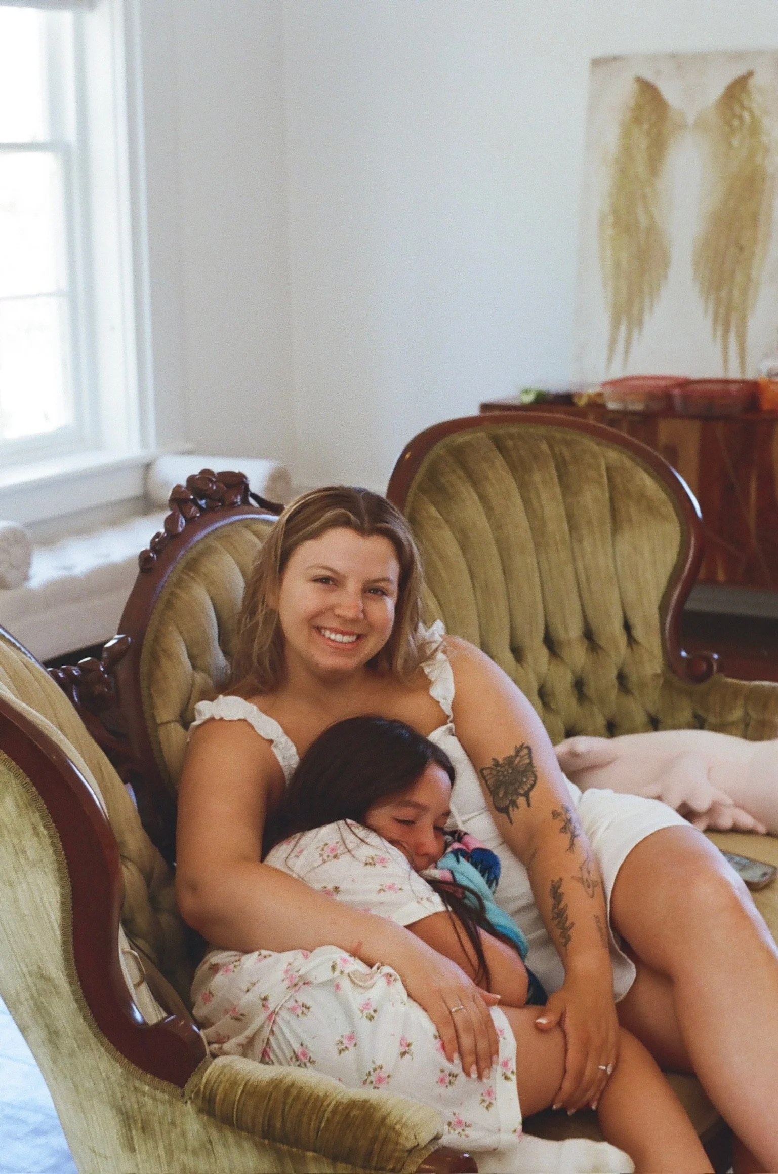 Two women and a young girl resting together on vintage upholstered chairs in a bright living room with a window and wall art.