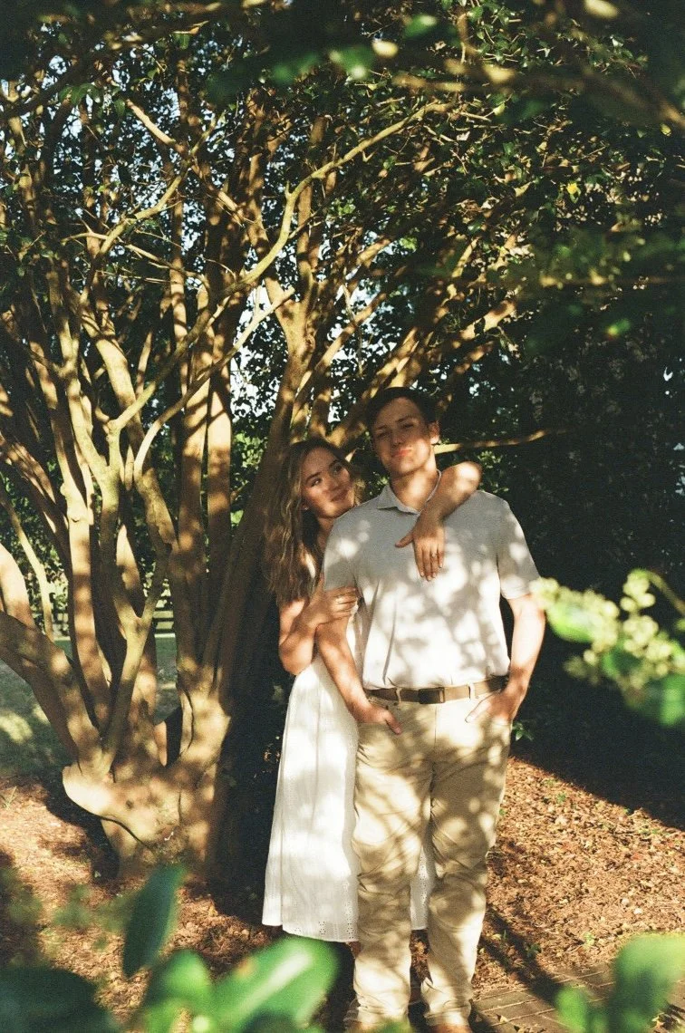 A young woman and young man stand close together under a large tree with twisted branches. The woman has long, wavy hair and is wearing a white dress, while the man is dressed in a light-colored polo shirt and khakis. They are posing for a photo outd
