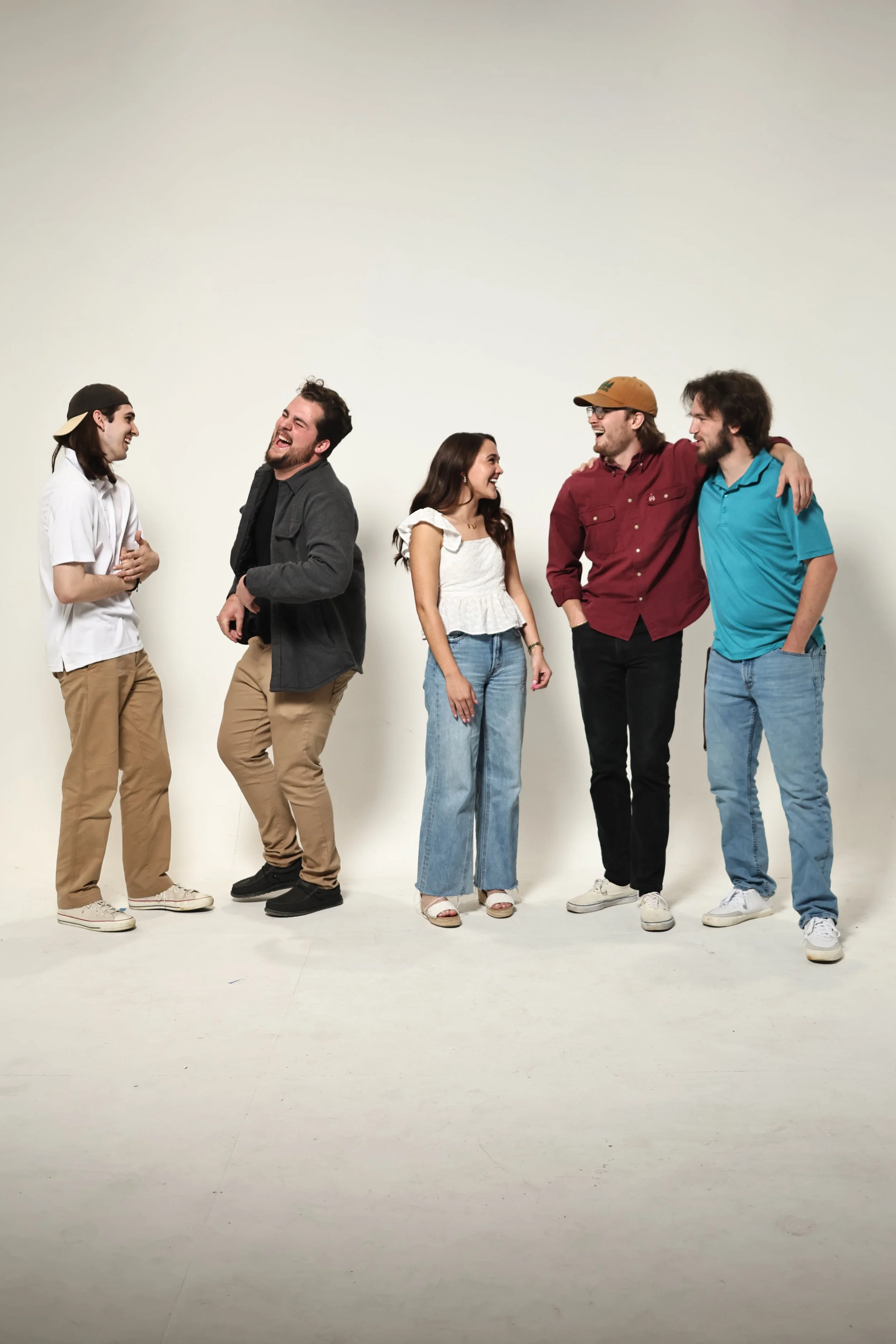 Six young adults standing together laughing and talking in a studio with a plain white background.