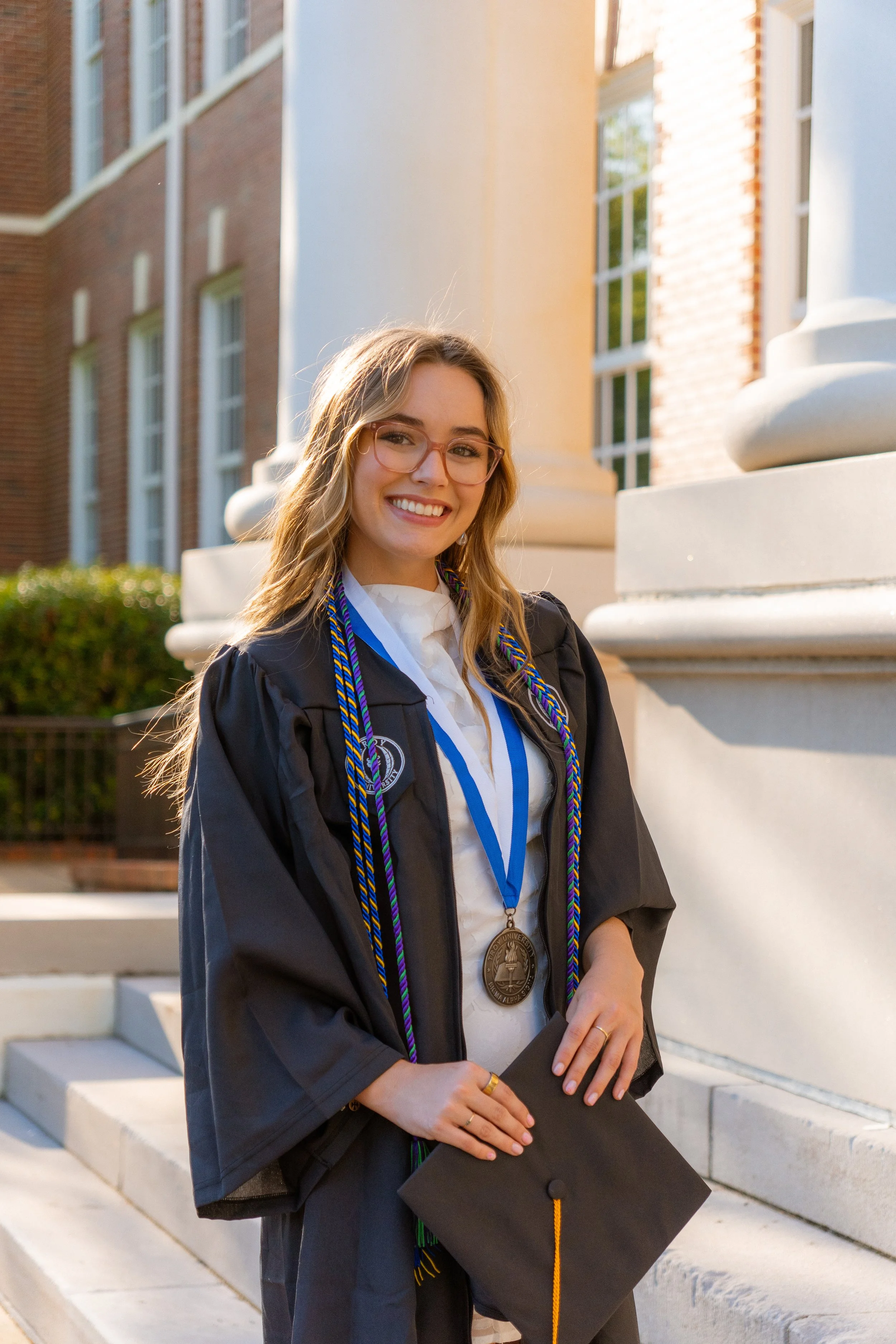 A young woman in graduation gown and cap standing outside on steps, smiling, wearing medals and cords, holding diploma, in front of a brick and stone building.