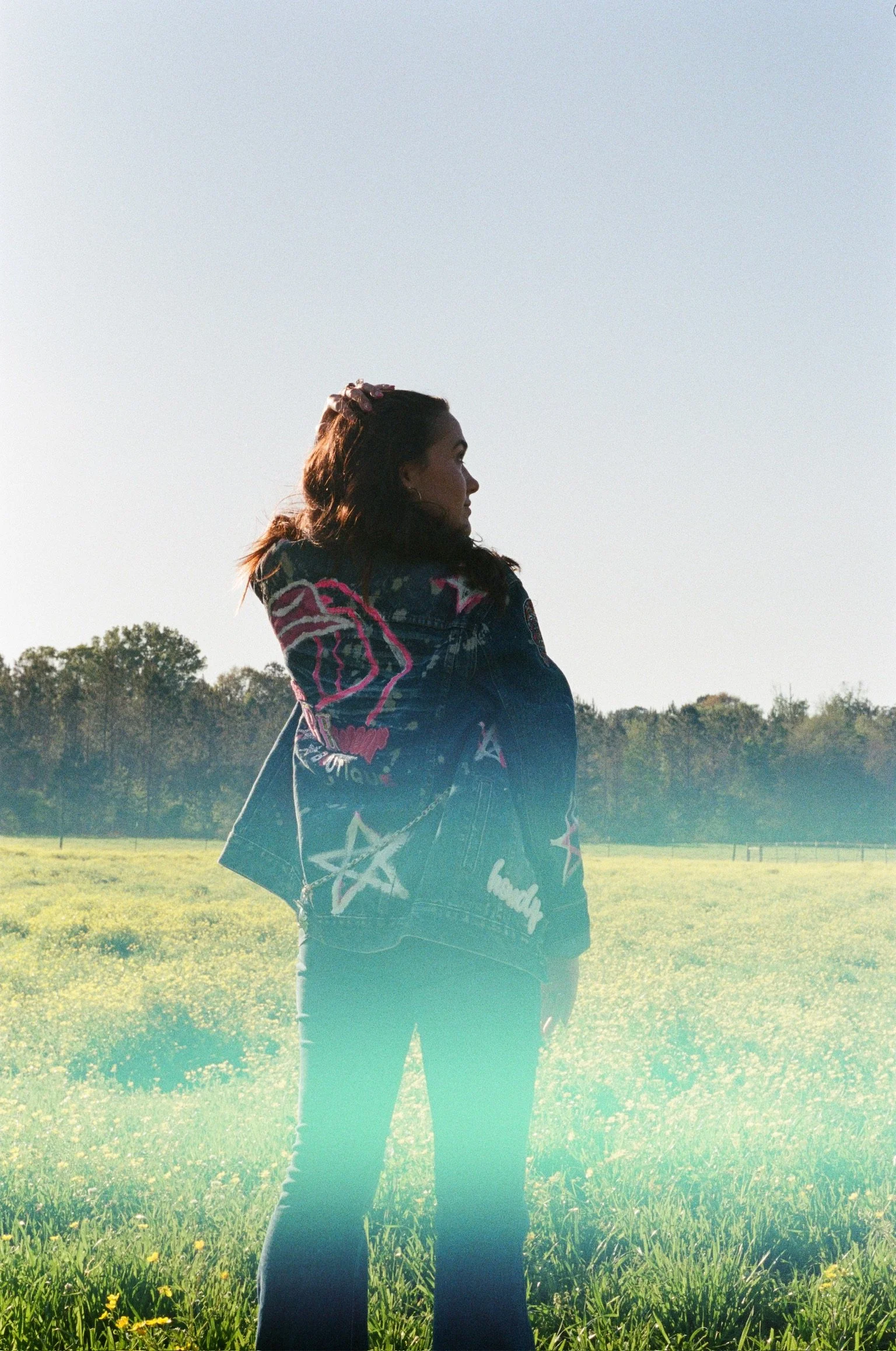A woman with brown hair standing outdoors in a grassy field, facing sideways with trees and a clear sky in the background. She is wearing a dark jacket and jeans.