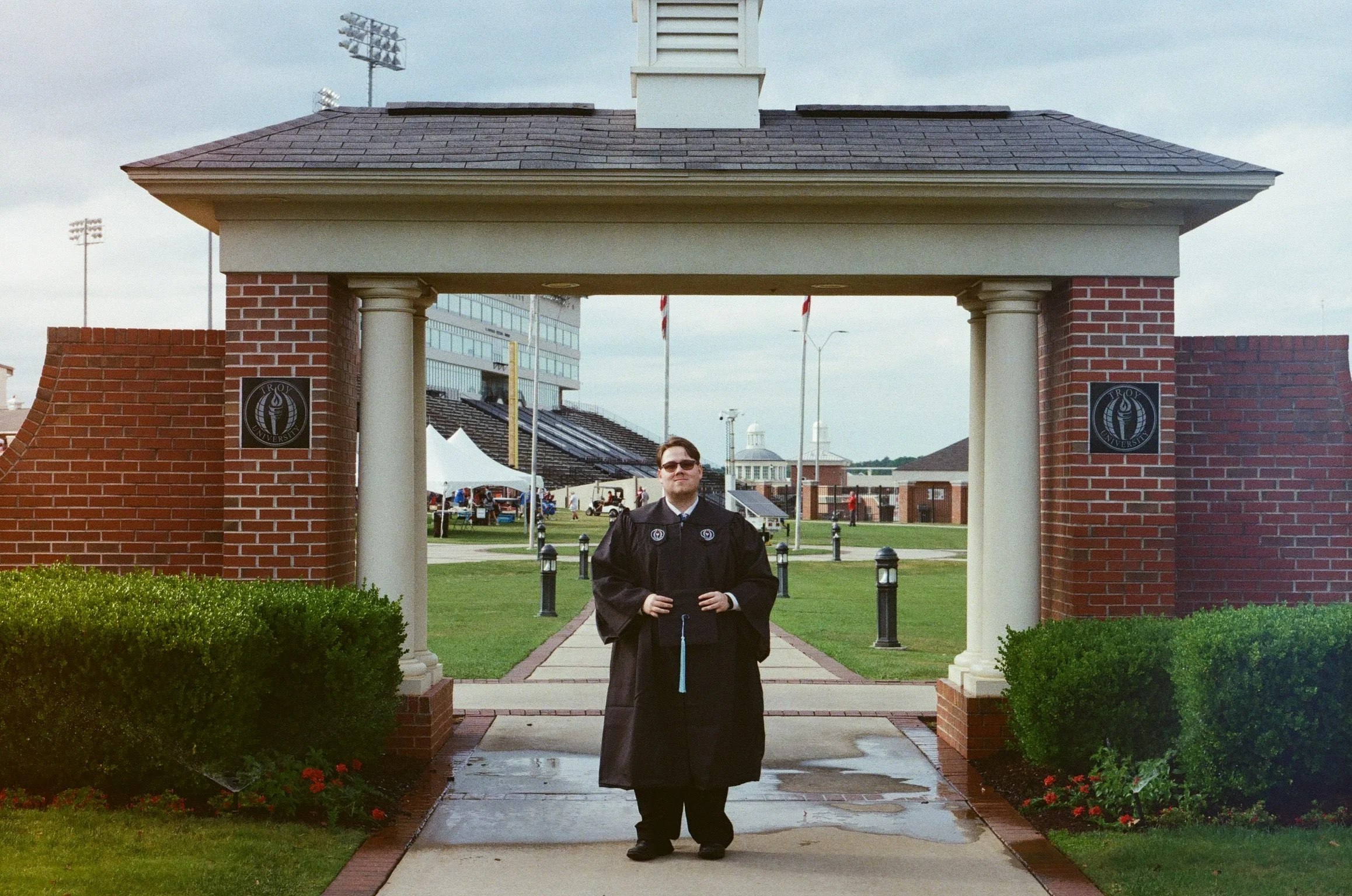 A person in a graduation gown standing under a decorative archway on a university campus with a football stadium in the background.