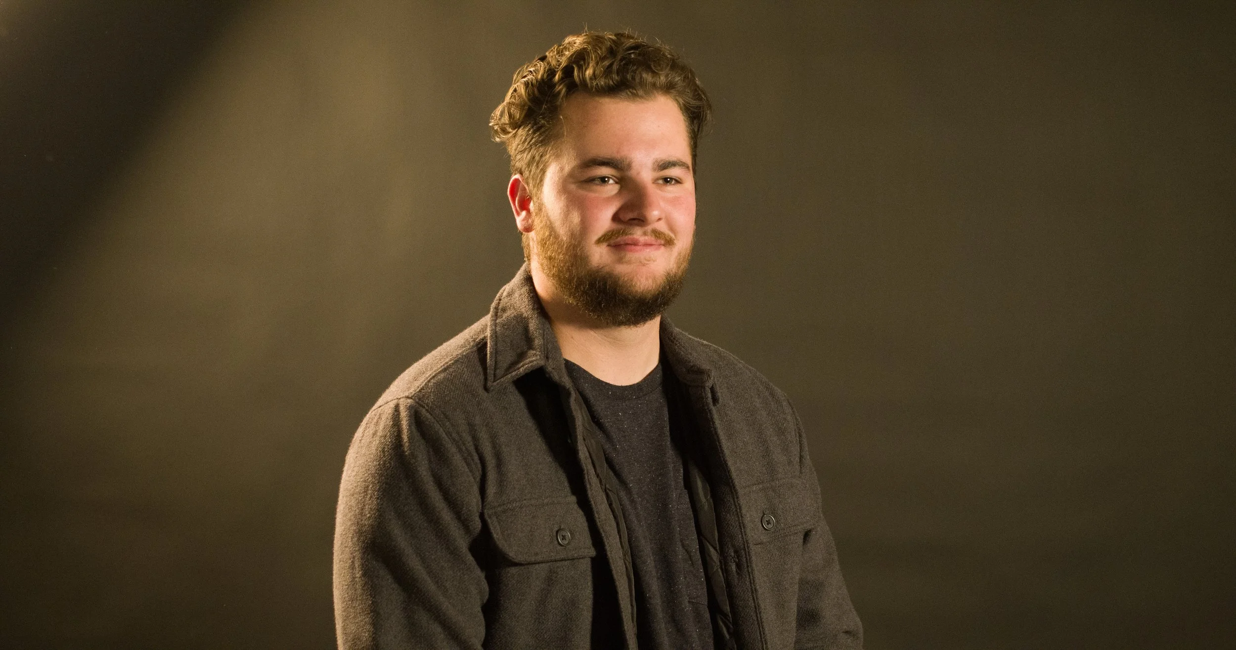 A young man with a beard and curly hair smiling, wearing a dark jacket over a t-shirt, standing against a plain background.