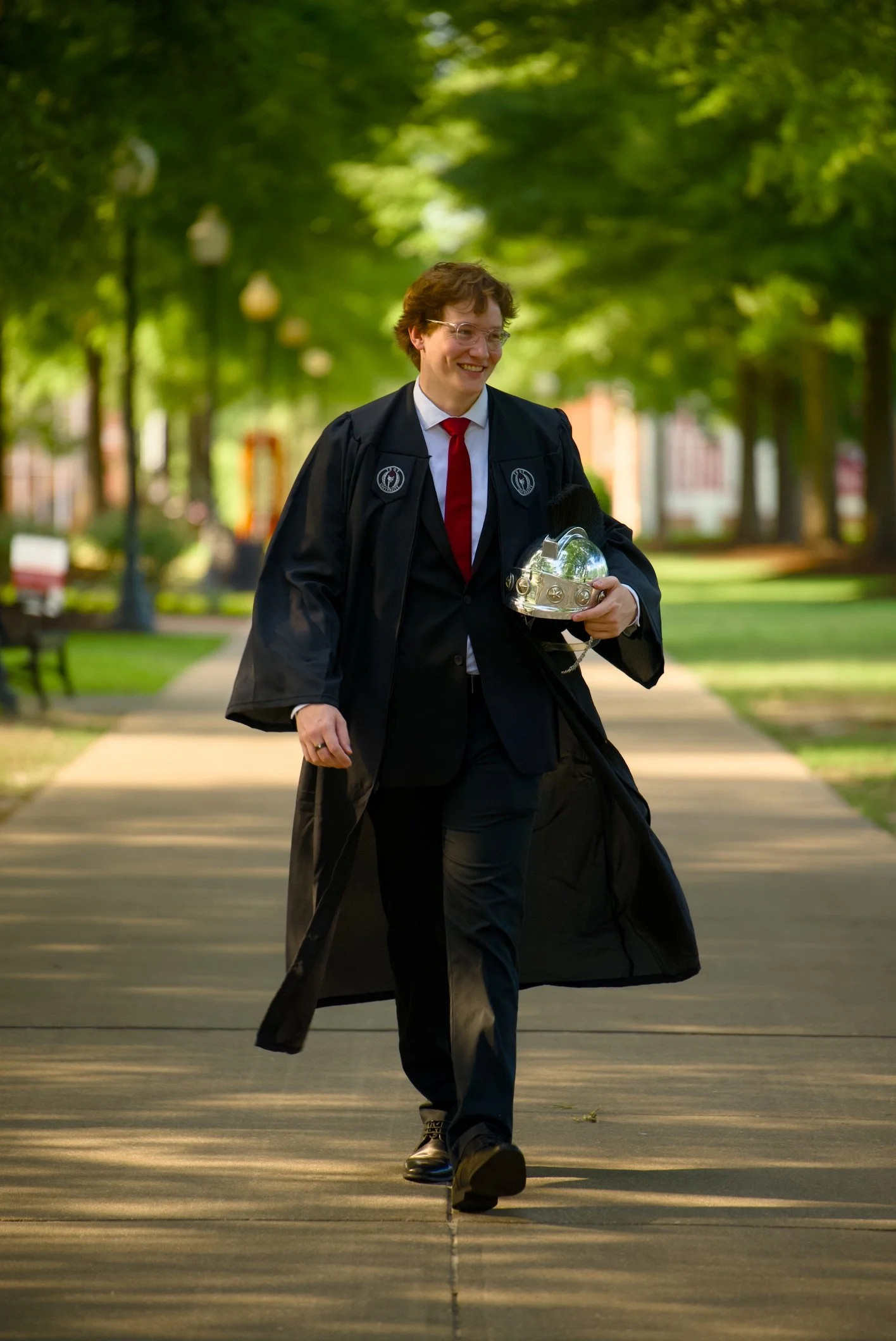 A person in academic regalia walking outdoors on a paved path, holding a shiny silver helmet, with green trees and lanterns in the background.