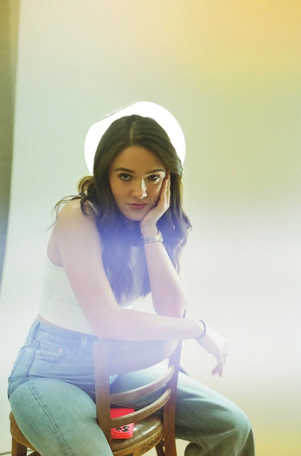 Young woman with long wavy brown hair sitting on a chair, resting her head in her hand, looking directly at camera, wearing a white tank top, light blue jeans, and a watch, with a bright light behind her.