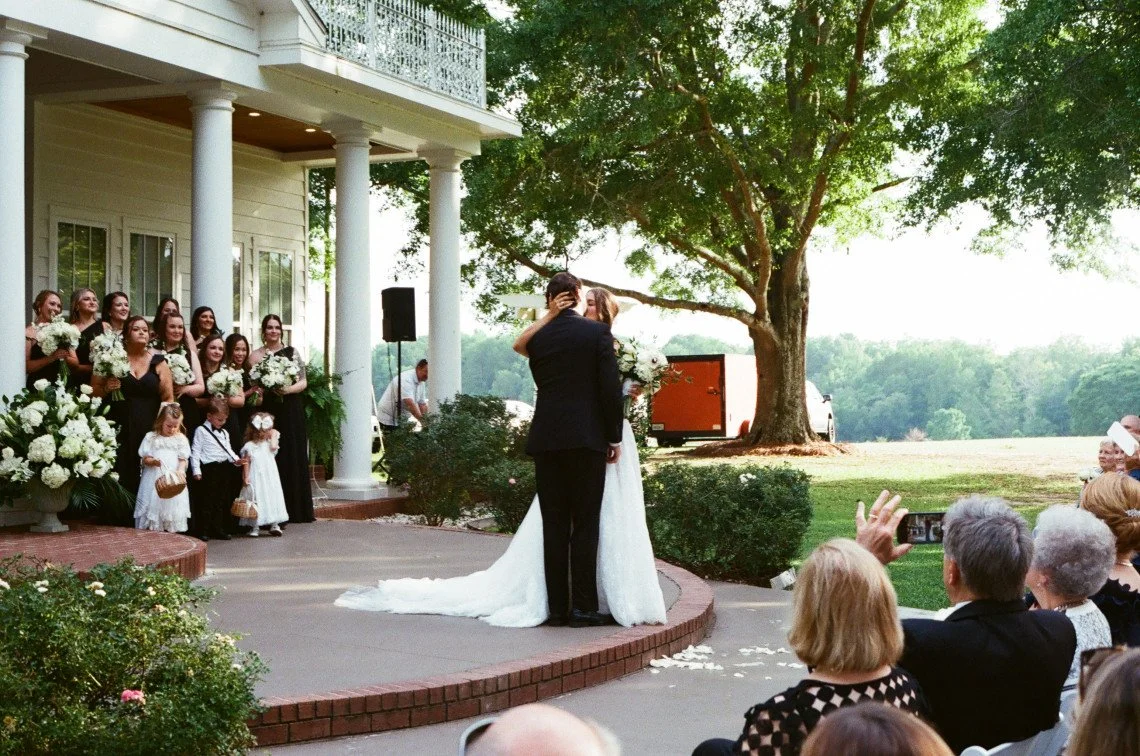 A couple shares a kiss during their outdoor wedding ceremony on a brick platform, with guests seated nearby and a wedding party standing to one side, all under a large tree on a sunny day.