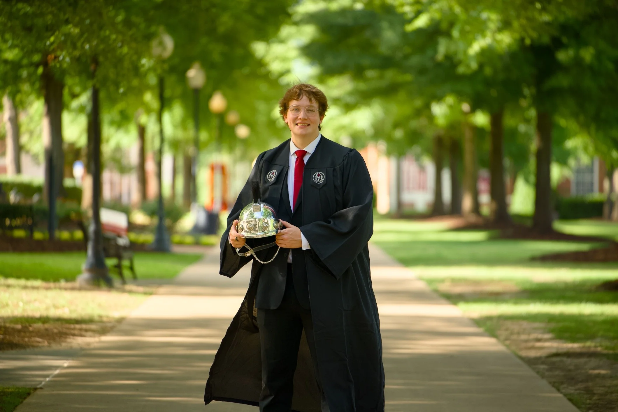 A young man in a graduation gown and red tie holding a silver graduation cap, standing on a sidewalk in a park with green trees and lamps.