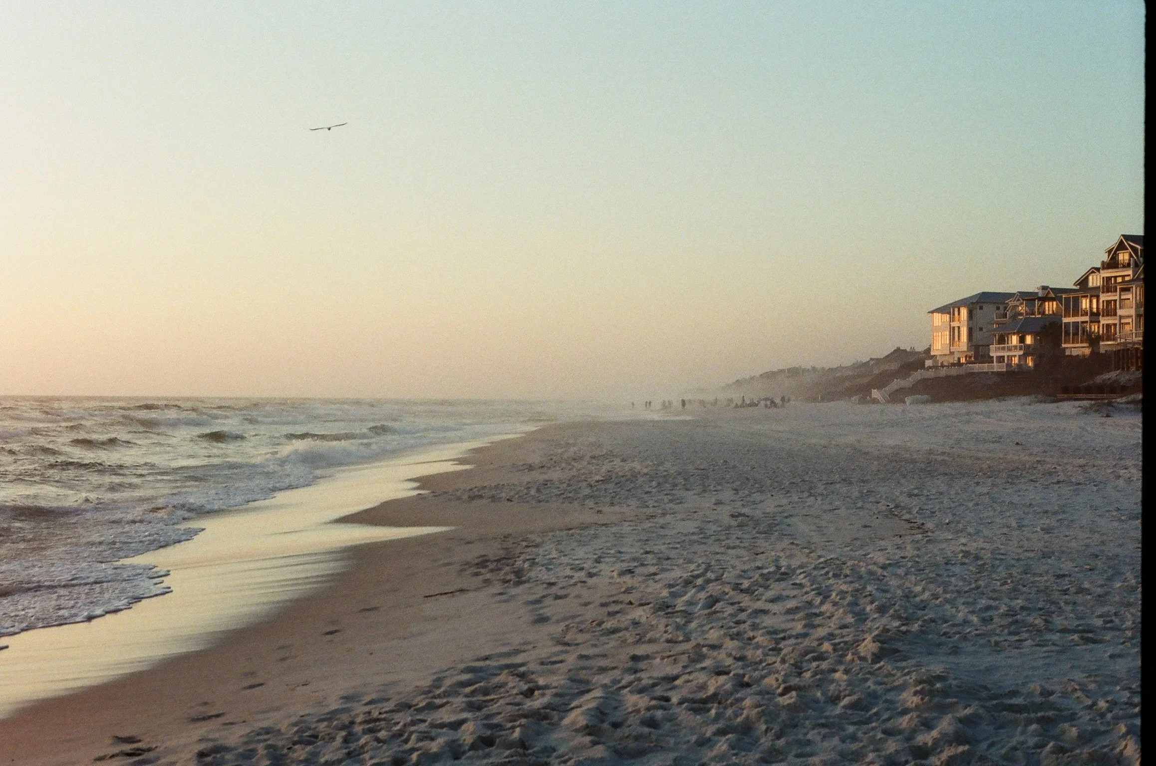 A beach during sunset or sunrise with waves approaching the sandy shore, multi-story houses on a cliff on the right, and a plane flying in the sky overhead.