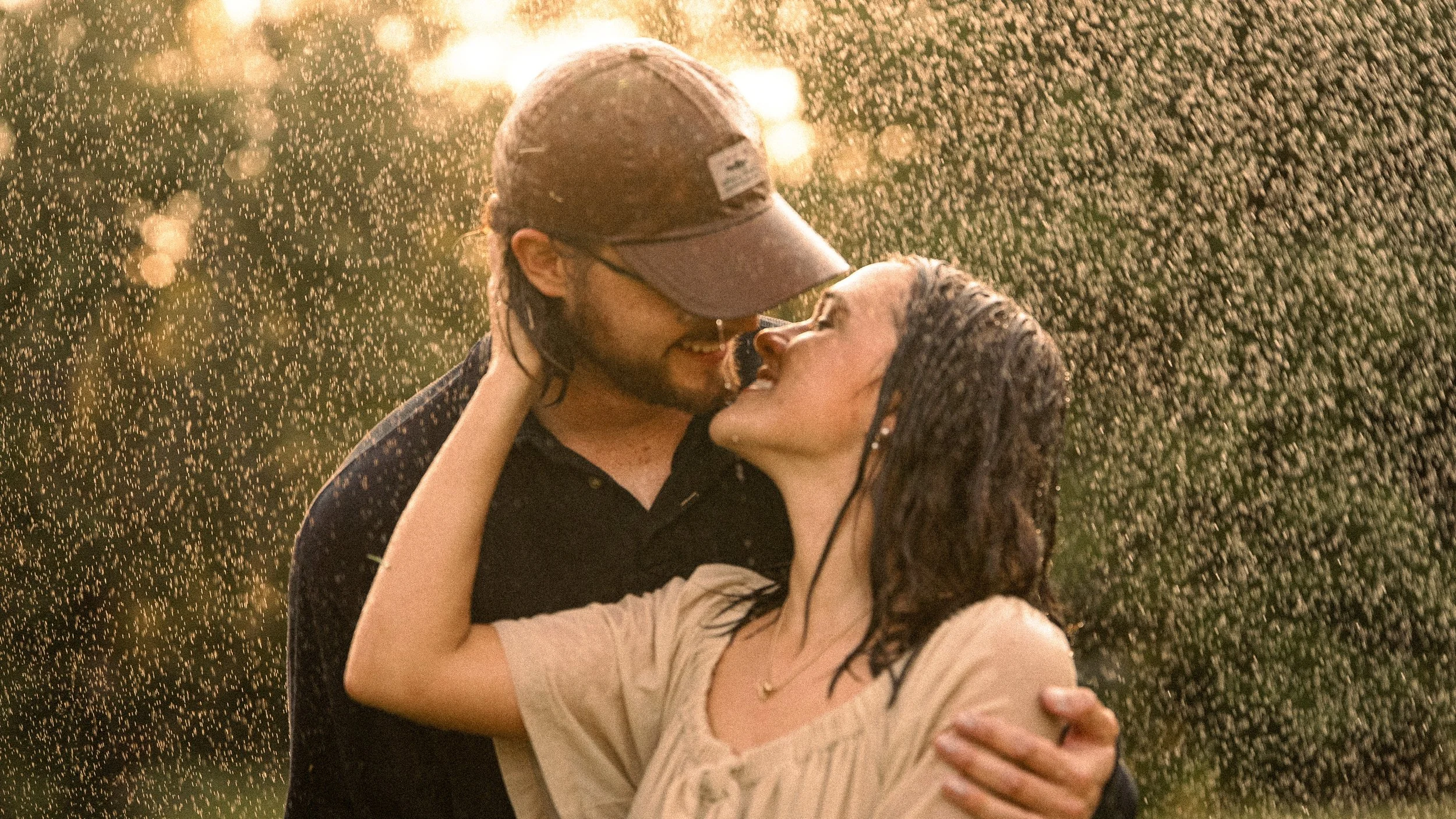 A couple is dancing and smiling in a rain shower during sunset, with water droplets illuminated by the golden light.