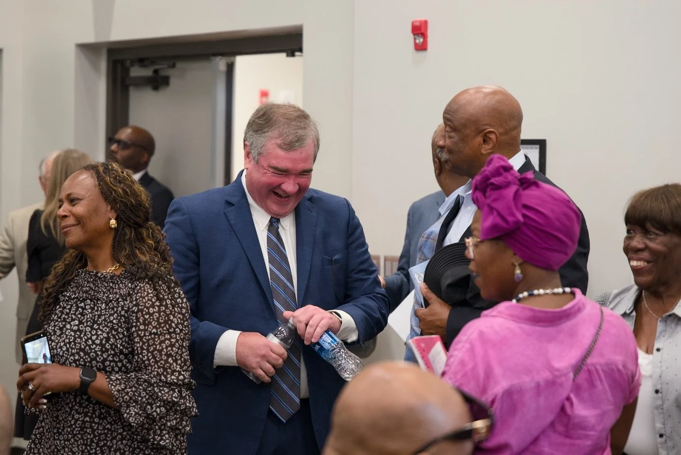 Group of diverse people in a celebratory or social setting, smiling and engaging with each other, with some holding items like water bottles, a phone, and a hat.
