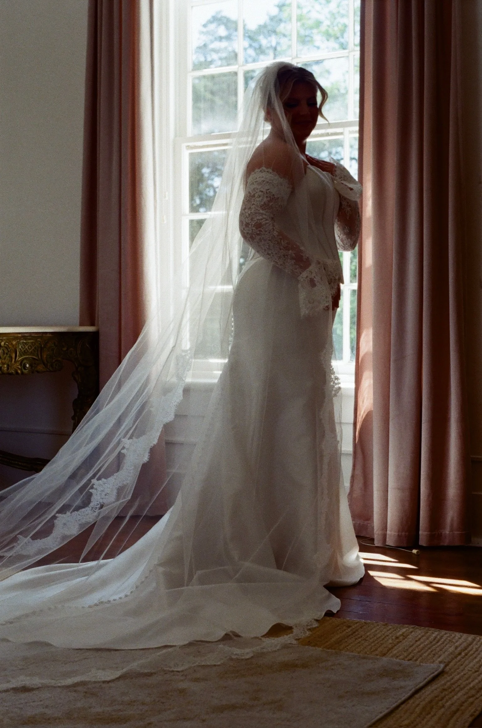A bride standing indoors near a window, wearing a lace wedding dress with a long veil, with sunlight coming through the window behind her.