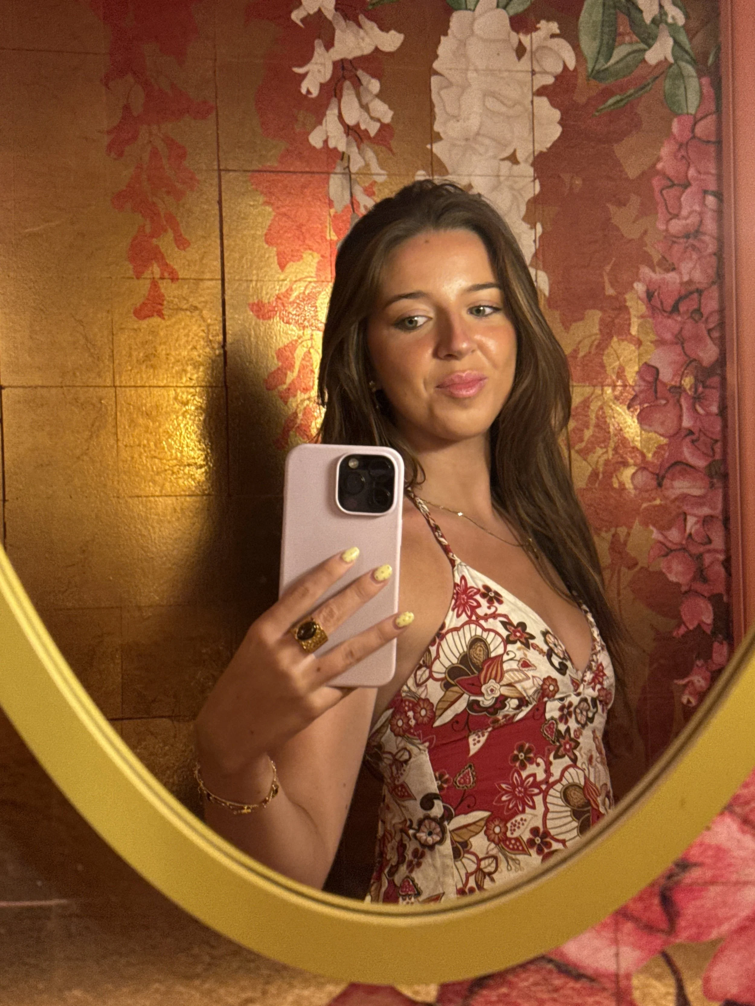 A young woman with brown hair taking a selfie in front of a decorative wall with floral patterns. She is wearing a dress with a red, white, and brown floral design, and has a ring on her finger, a bracelet, and earrings. The photo is captured in a mirror with a yellow frame.