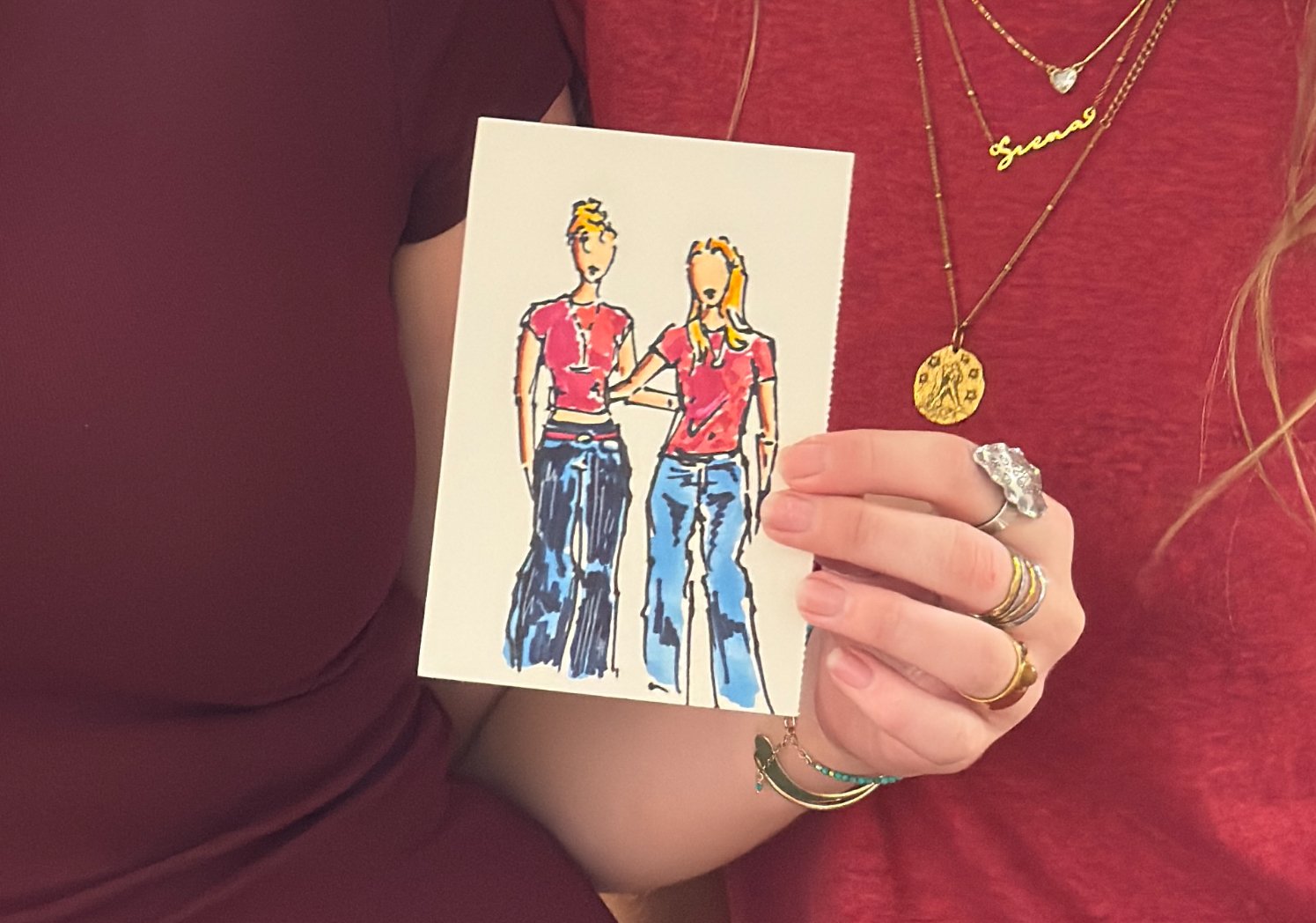 Person holding a drawing of two women with their arms around each other's shoulders. One woman has short hair, and the other has long hair. Both are wearing red tops and blue jeans.