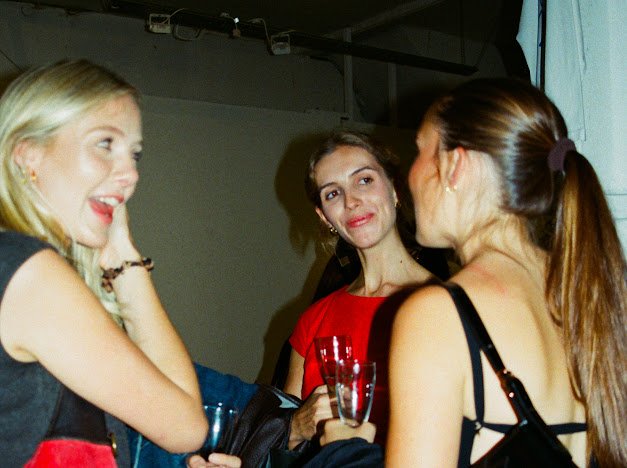 Three women at a social gathering, talking and smiling. One woman has blonde hair and is wearing a black dress with red accents, another has brown hair and is wearing a red top, and the third has brown hair tied back and is wearing a black dress. One woman is holding a glass of drink.