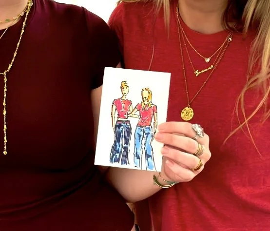 Two women holding a sketch of two women wearing red shirts and blue jeans.