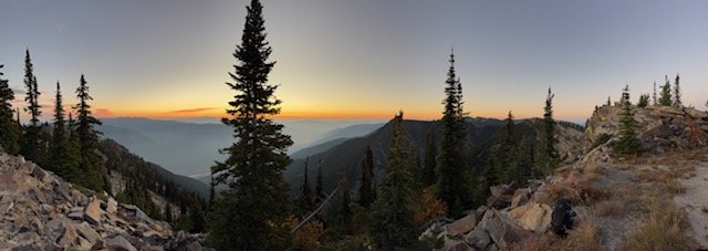 Mountain landscape at sunset with tall pine trees and rocky terrain.