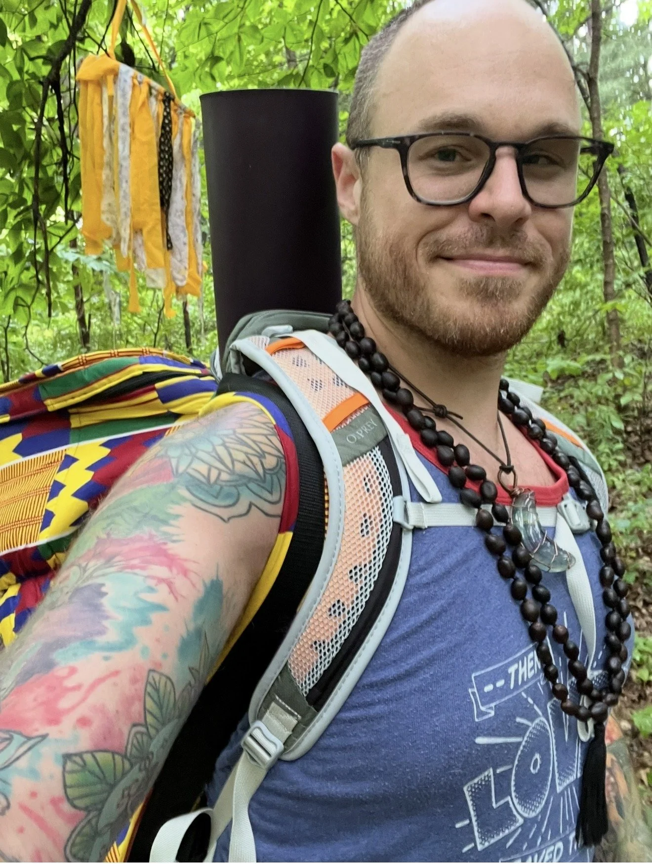 A smiling man with glasses, tattoos on his arm, and beaded necklaces taking a selfie in a forest. He's wearing a blue T-shirt, a backpack with a rolled-up mat attached, and a yellow and multicolored strap. There is a yellow cloth with black strings hanging from a tree behind him.
