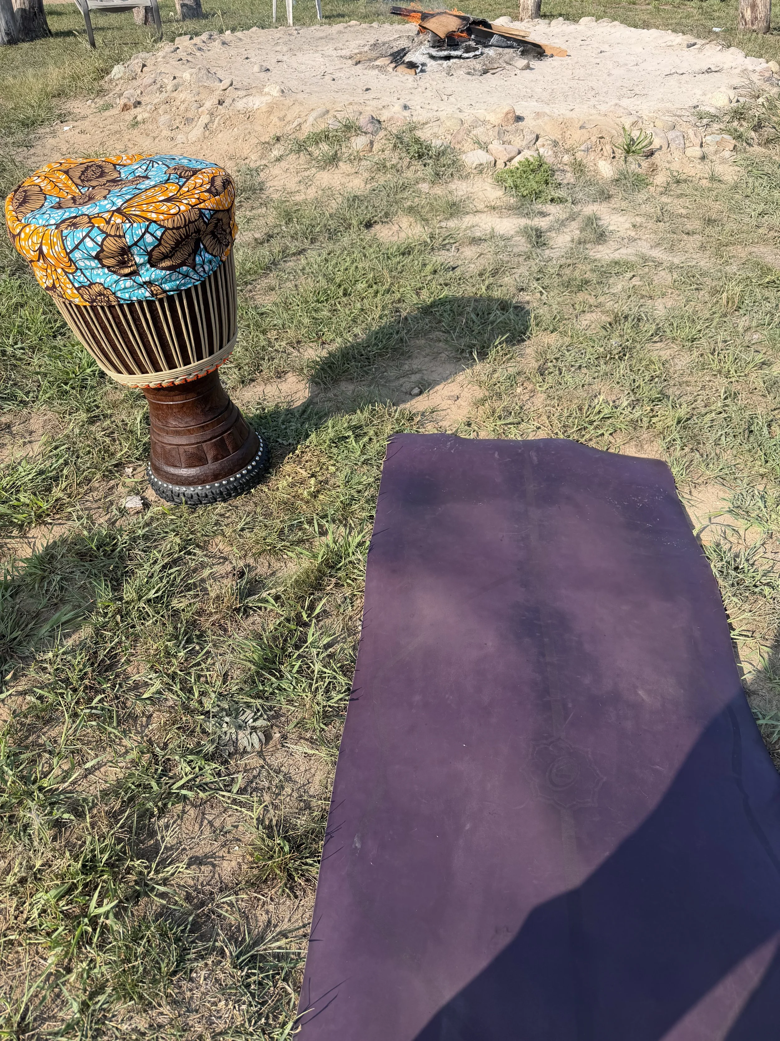 A drum with a colorful butterfly pattern head cover, placed on grass near a worn purple yoga mat and a fire pit with ashes and smoldering wood.