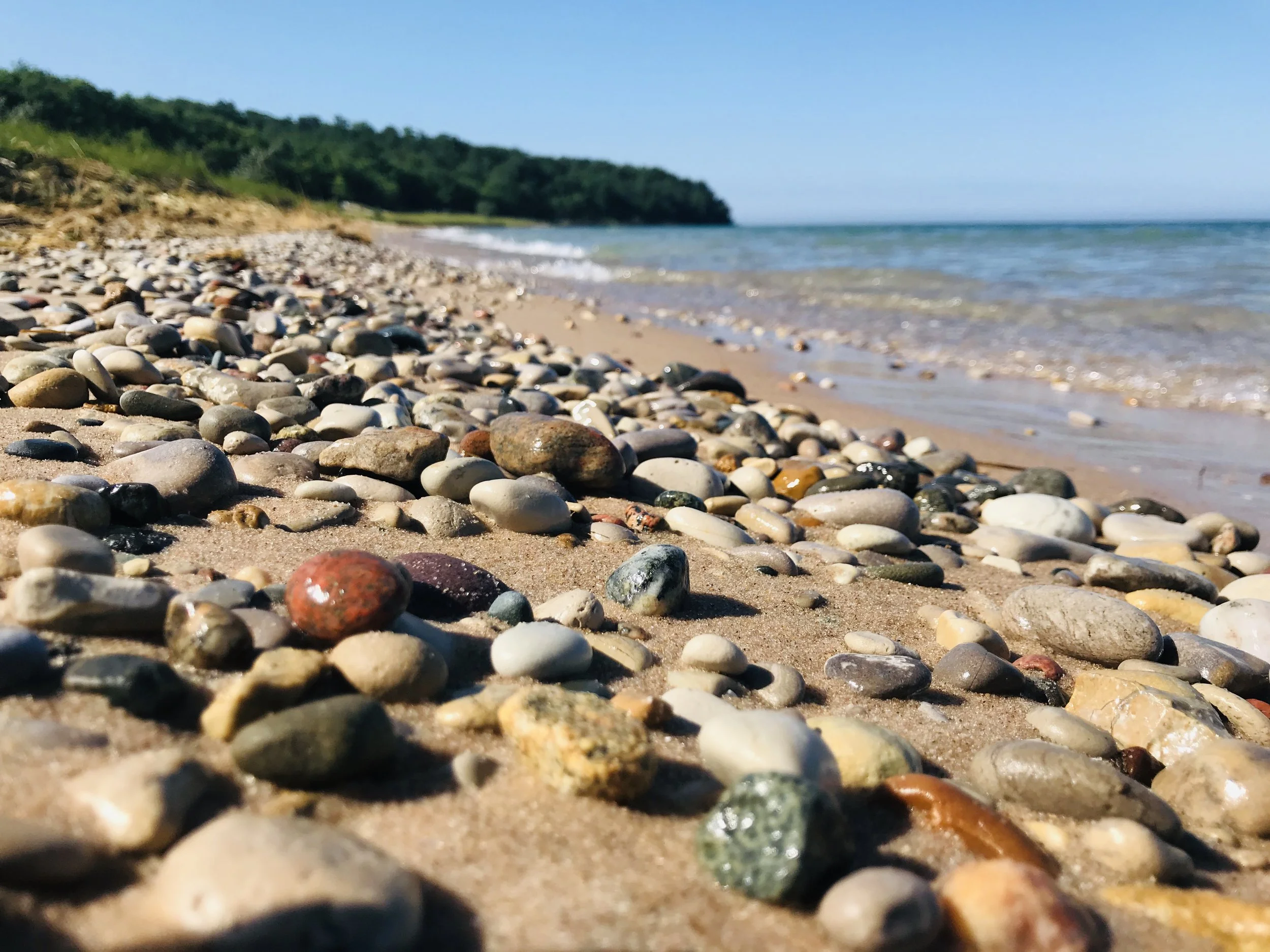 Close-up view of a pebble-covered beach with a forested hill in the background and a cloudy blue sky.