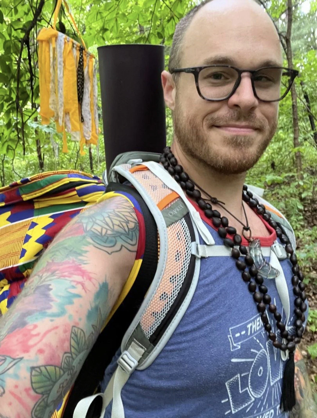 A smiling man with glasses, tattoos on his arm, wearing a blue shirt, hiking backpack with a yoga mat attached and a djembe in a lush green forest.