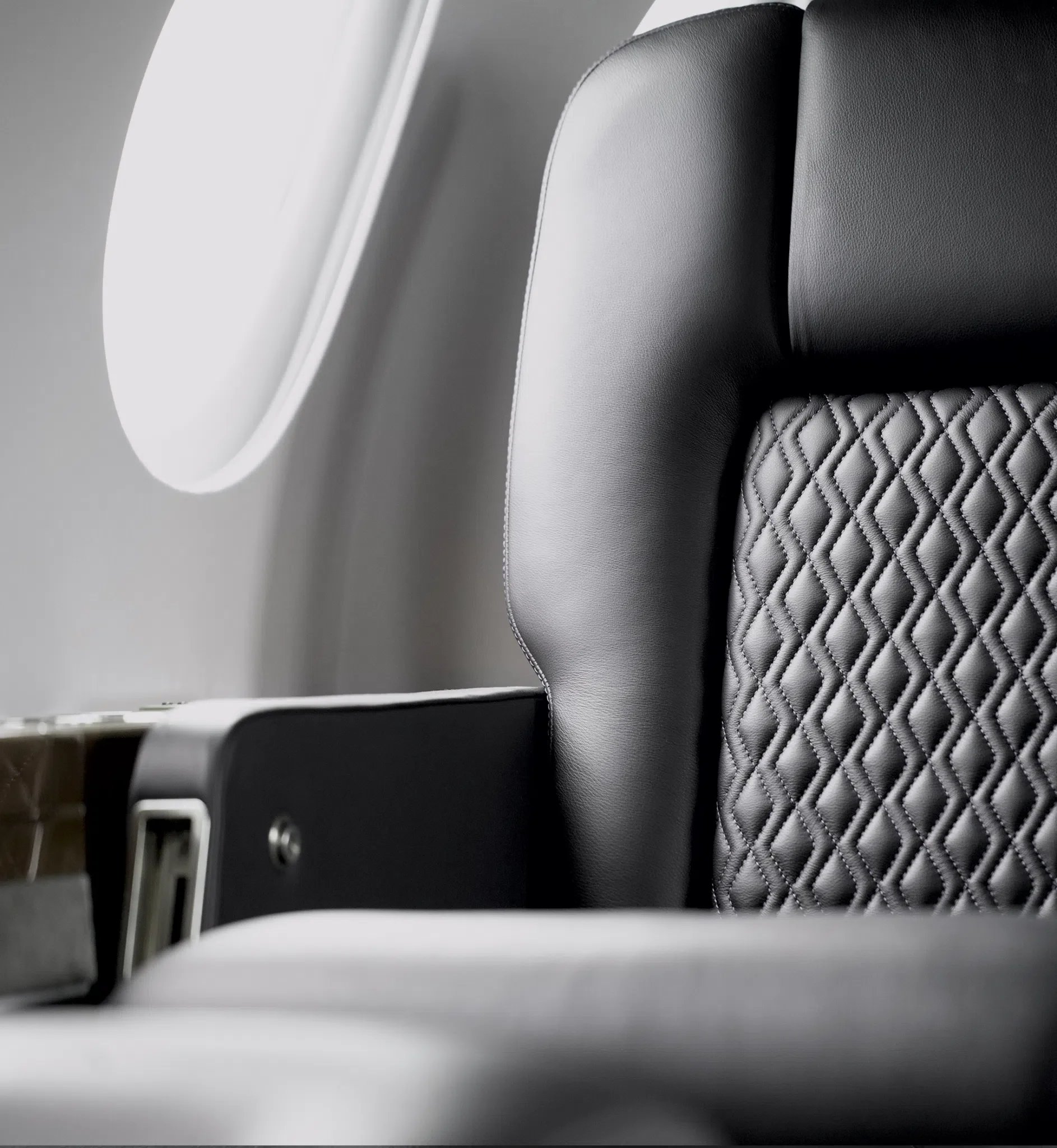 Nahaufnahme eines Flugzeugsitzes mit schwarzem Lederbezug und Quernähten, neben einem Fenster.