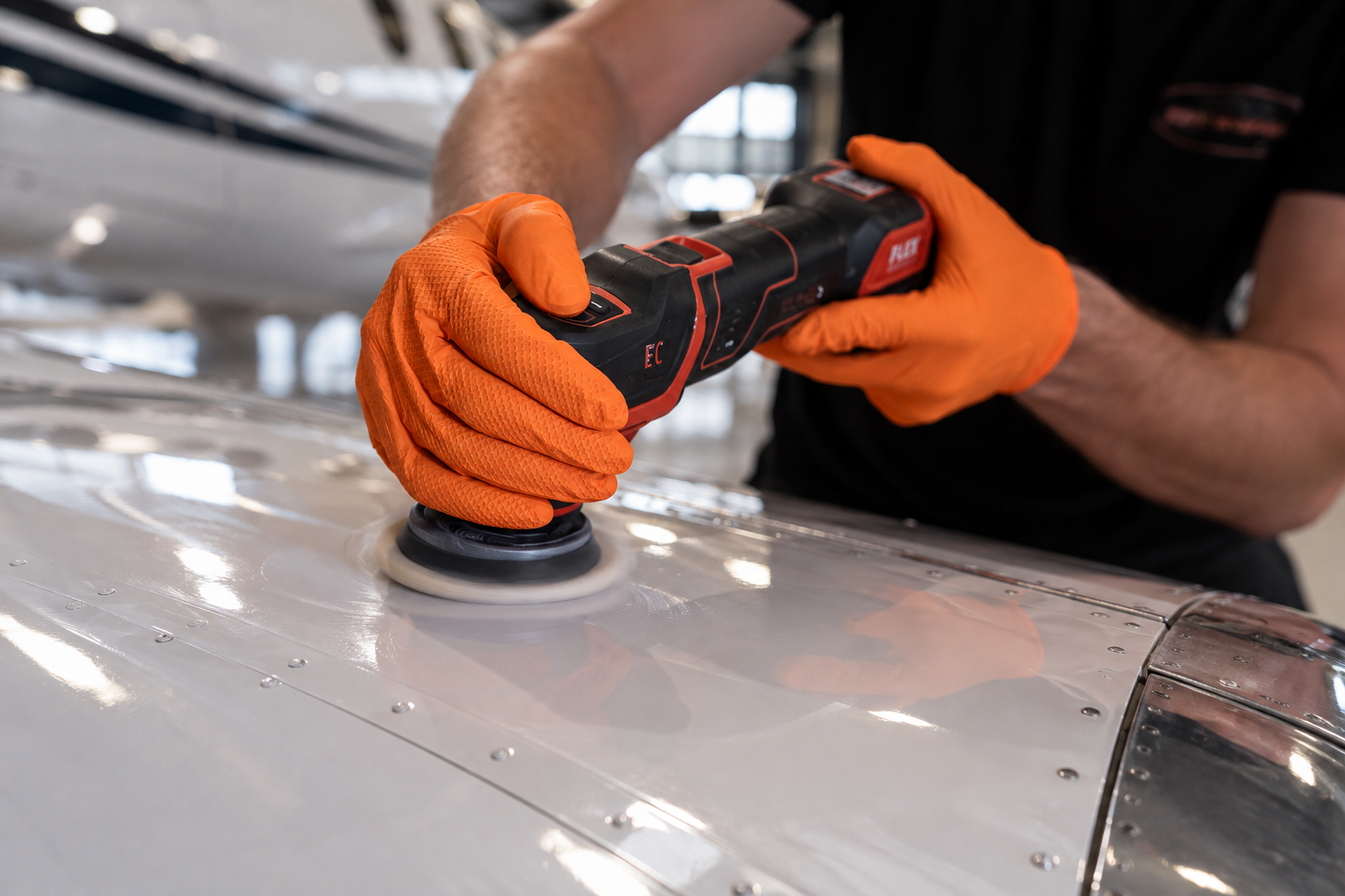 Person poliert eine Metallfläche mit einem elektrischen Poliergerät, trägt orange Handschuhe, in einer Werkstatt oder Flugzeughalle.