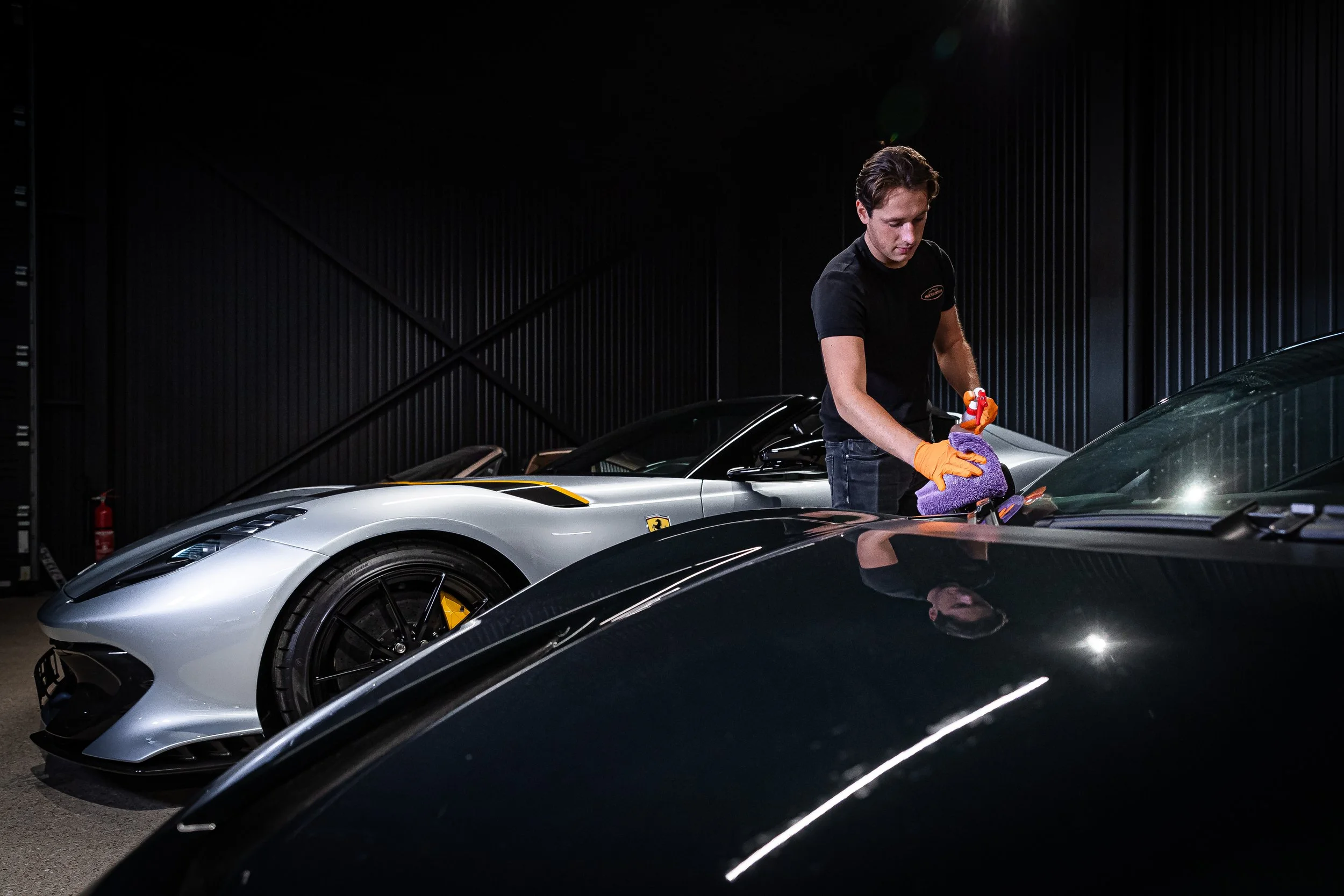 Ein Mann im schwarzen T-Shirt mit orangefarbenen Handschuhen putzt einen schwarzen Sportwagen in einer dunklen Garage, neben einem silbernen Lamborghini.