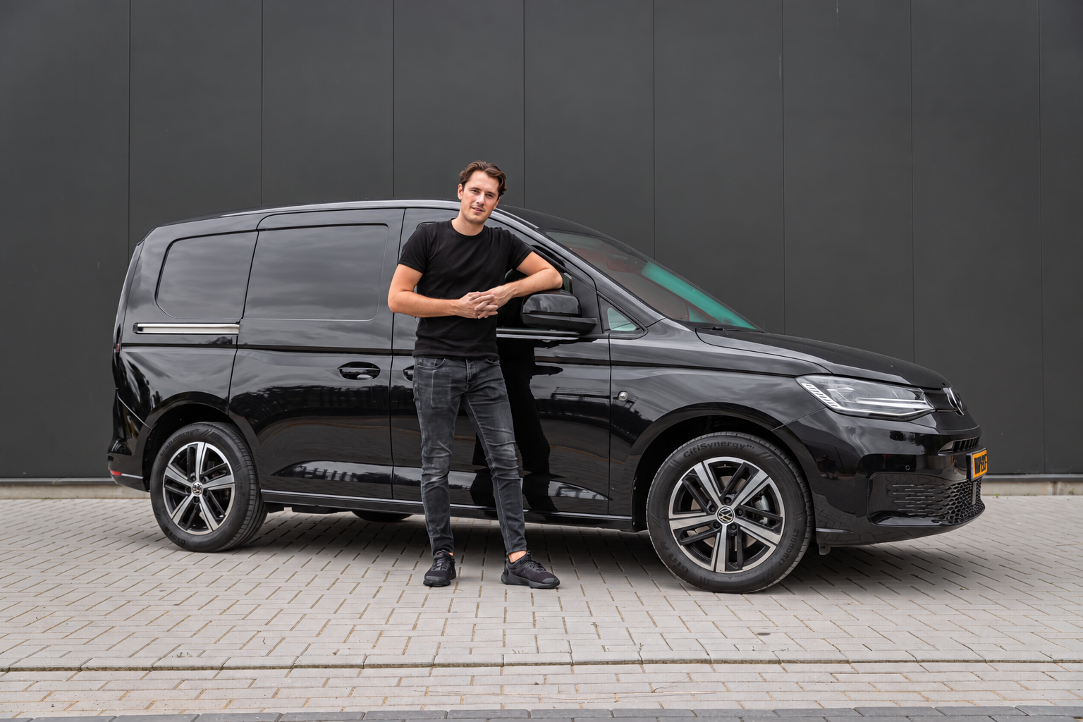 Ein junger Mann in schwarzem T-Shirt und Jeans steht neben einem schwarzen VW-Transporter vor einer dunklen Wand auf gepflastertem Boden.