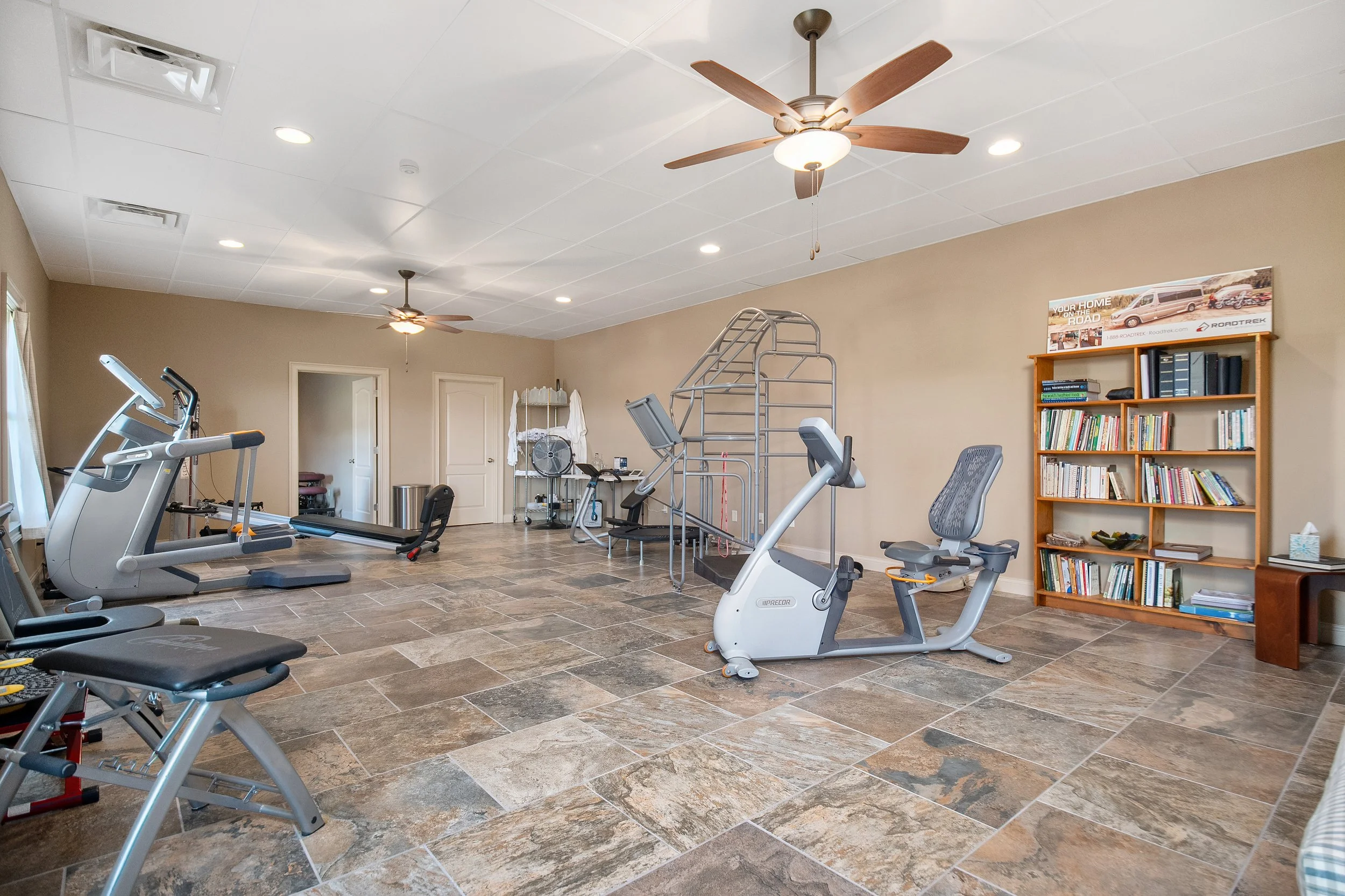 A basement gym room at the Ocoee River Estate in Benton, TN with exercise equipment including treadmills, stationary bikes, and a weight machine, with a bookshelf on the right side and ceiling fans and lights on the ceiling.