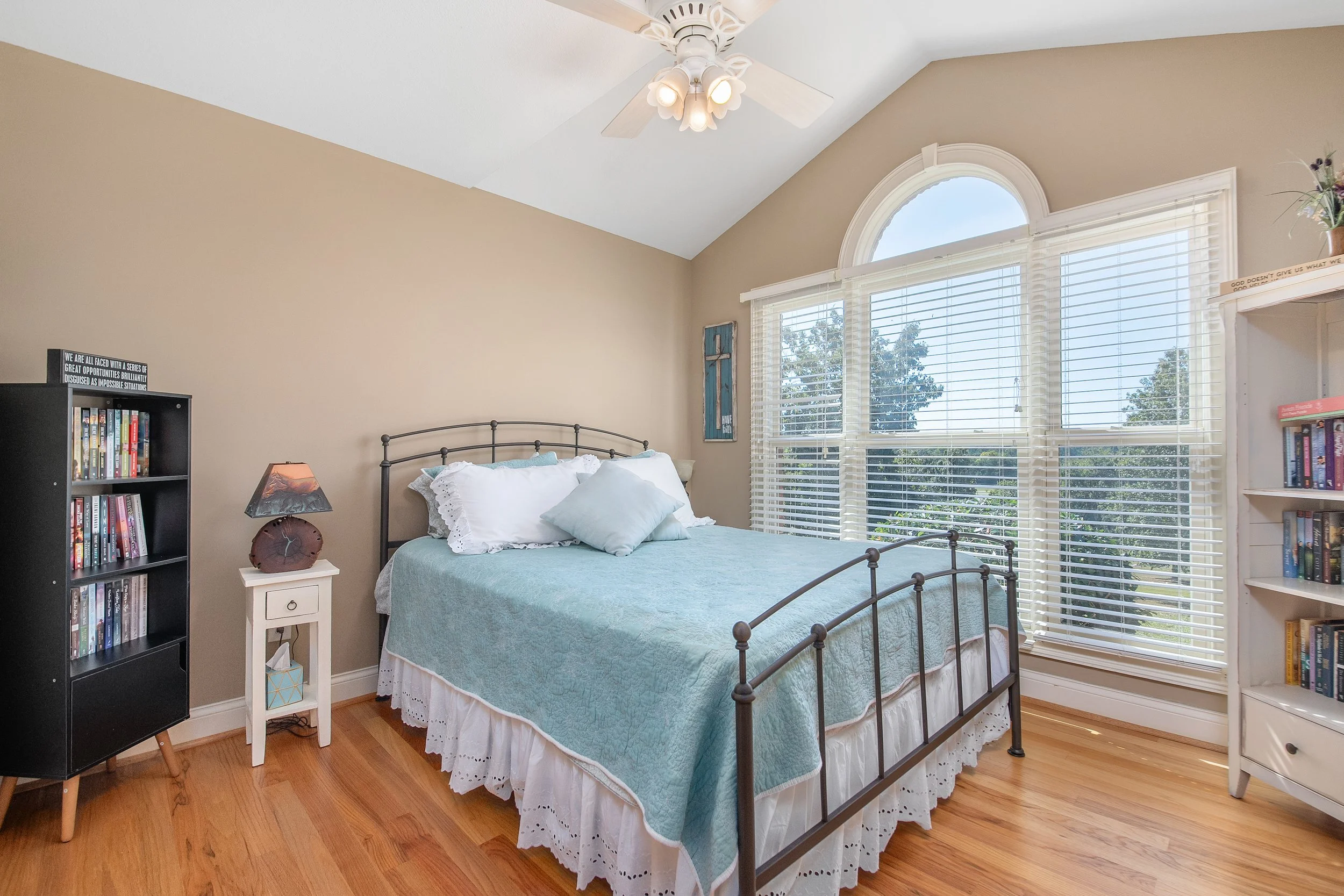 A bedroom with a metal-framed bed, white curtains covering large windows, and a ceiling fan with lights.
