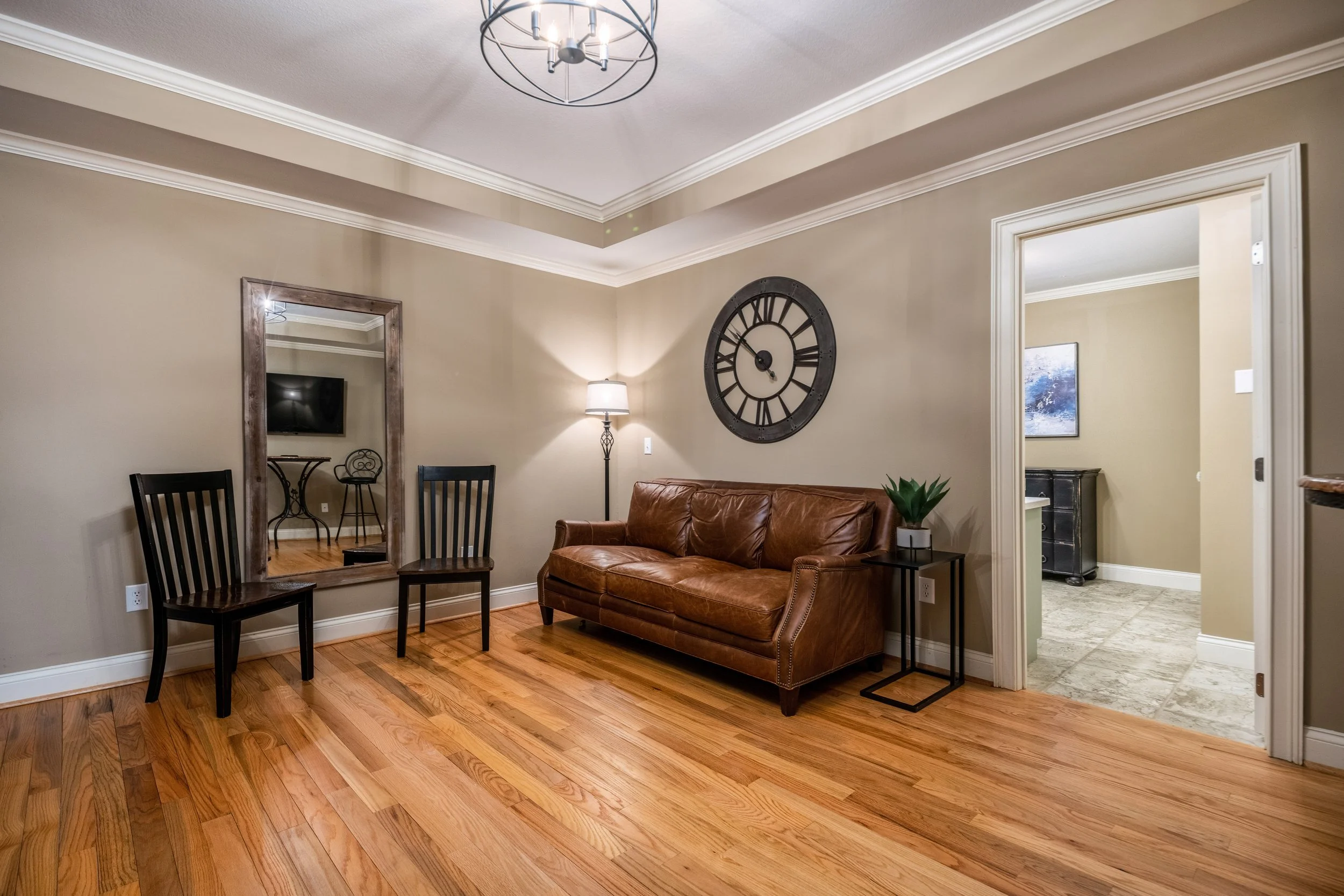 Living room with hardwood floors, a brown leather sofa, a black side table with a green plant, a standing lamp, a large wall clock, and a doorway leading to another room with a painting on the wall.