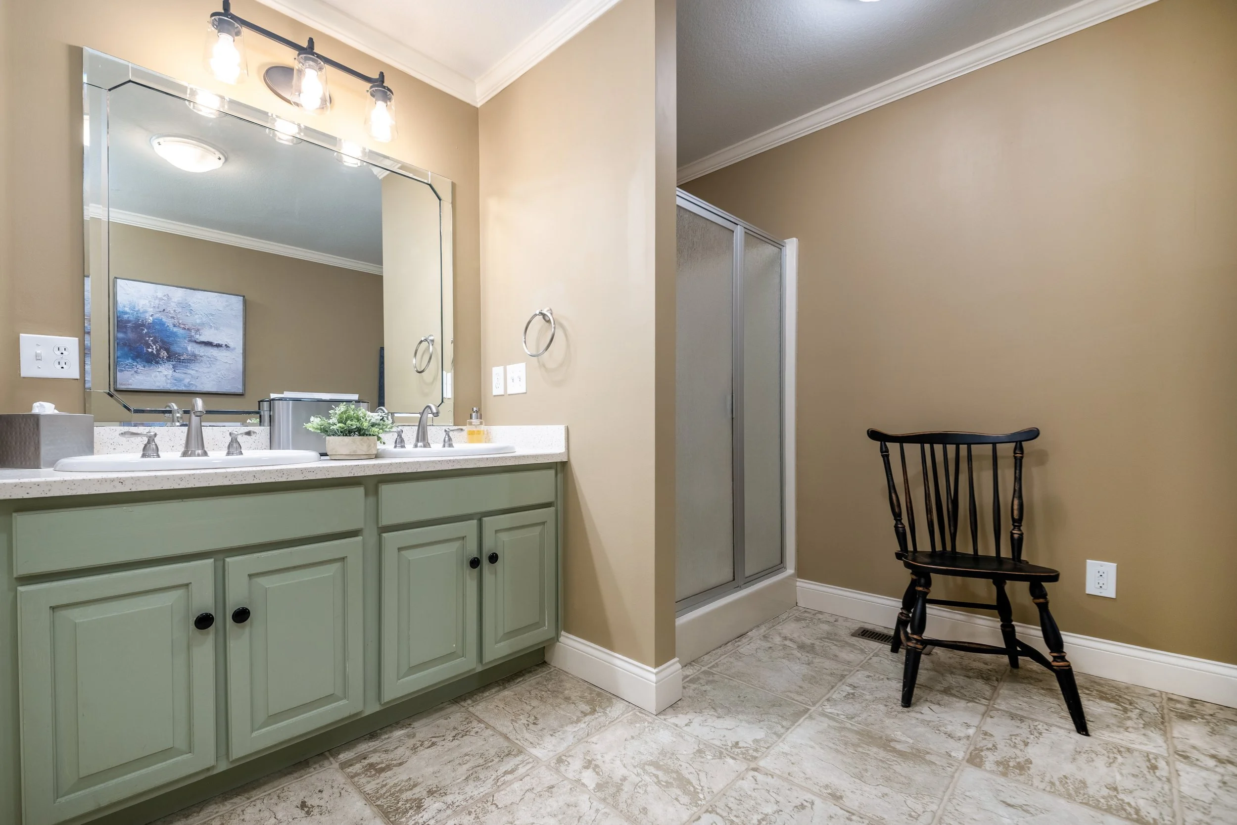 Bathroom with a green vanity, a large mirror, a light fixture with three bulbs, a white sink with silver fixtures, a small plant on the counter, a soap dispenser, a towel ring, a shower with frosted glass door, a beige wall, a black wooden chair, and