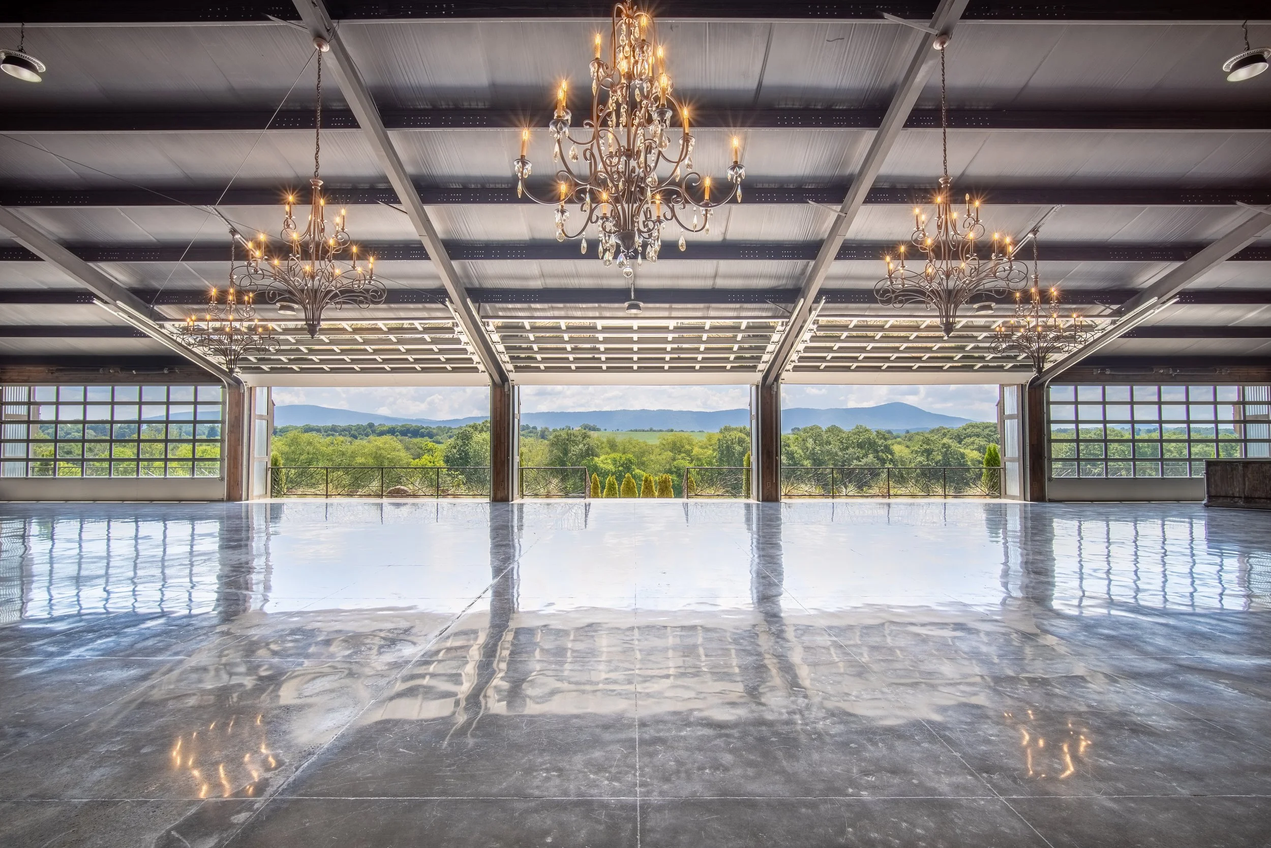 Elegant indoor space with hanging chandeliers, large open floor, and an expansive view of green trees and mountains through large windows.