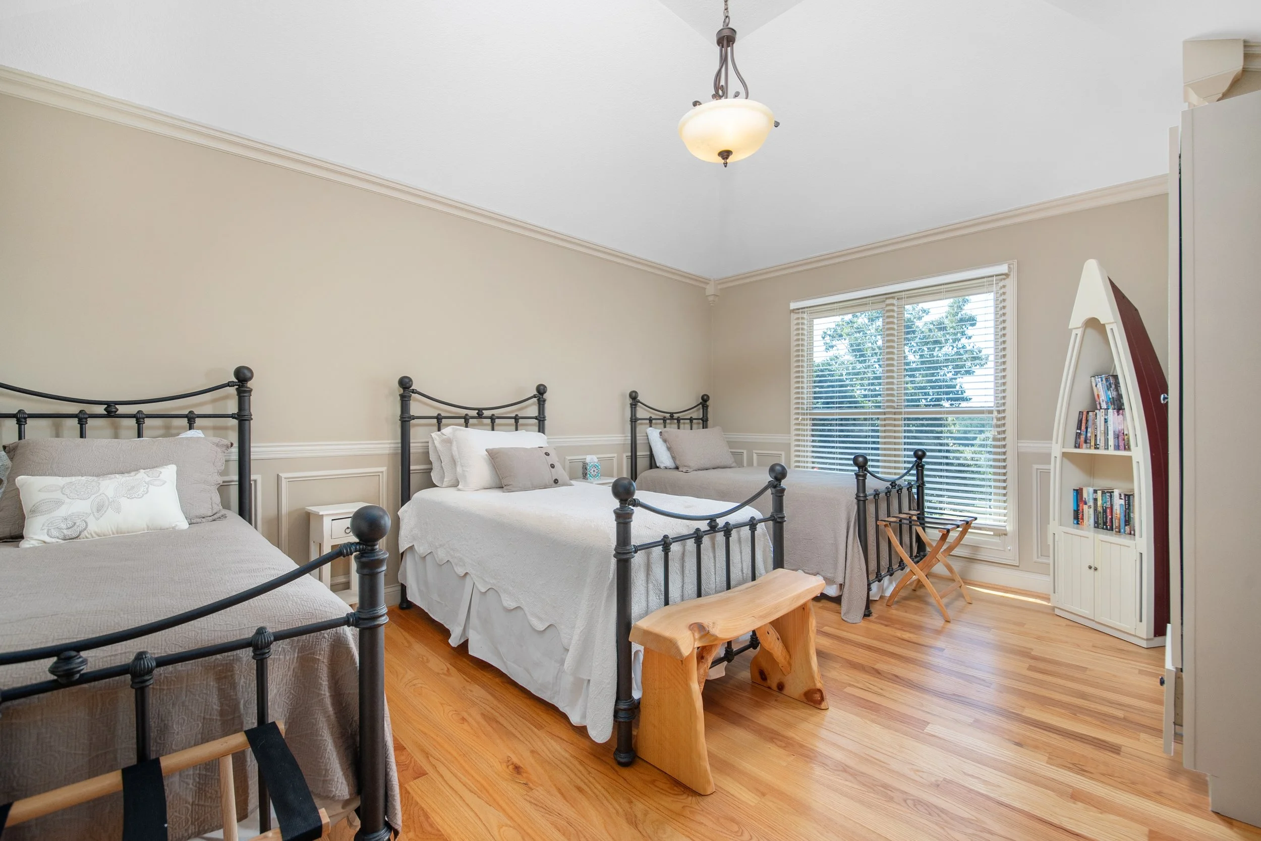 A bedroom with three beds featuring black metal frames, white and beige bedding, a window with blinds, and a wooden bench at the foot of one bed. There is also a louvered white bookshelf filled with books and a small wooden folding chair near the boo