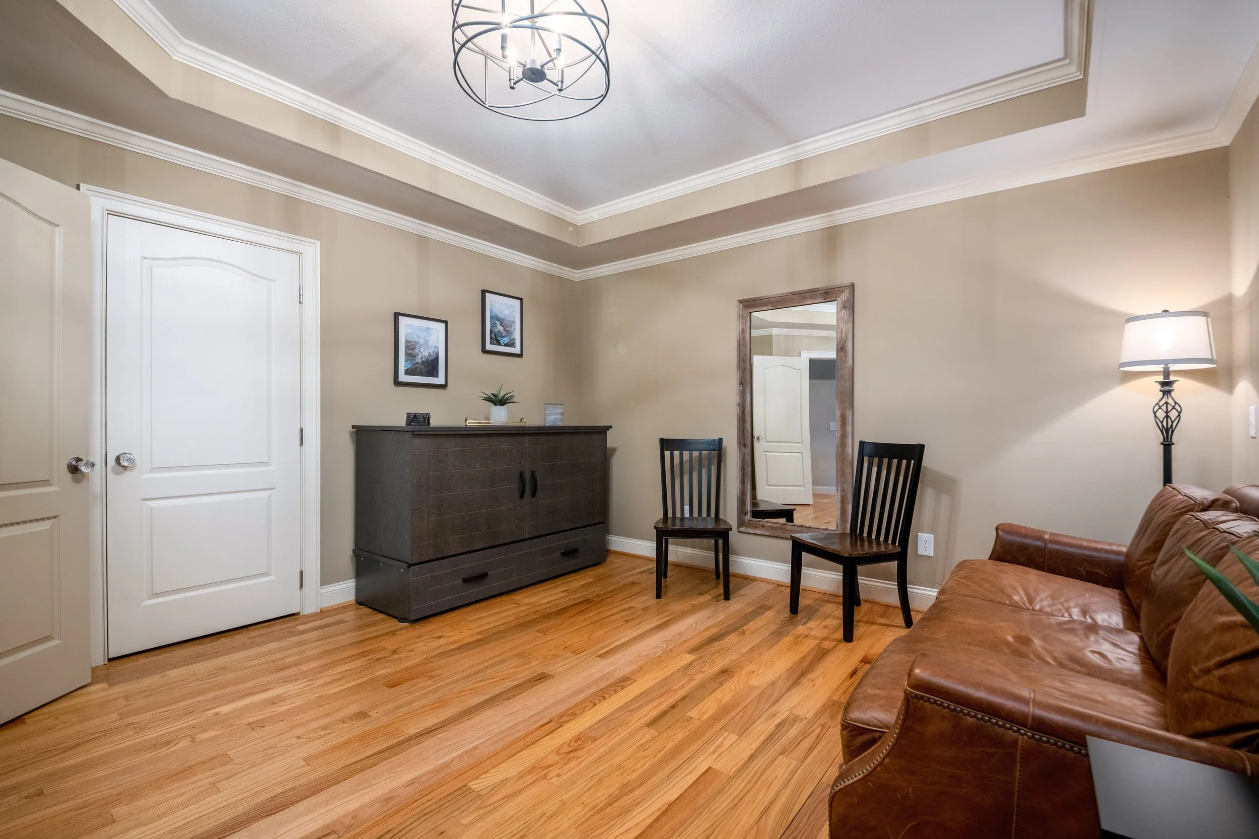 Interior of a bedroom at Ocoee River Estate in Tennessee with beige walls, hardwood floors, a brown leather couch, a black sideboard, and a tall mirror. There are two wooden chairs, a modern floor lamp, and framed art on the wall.