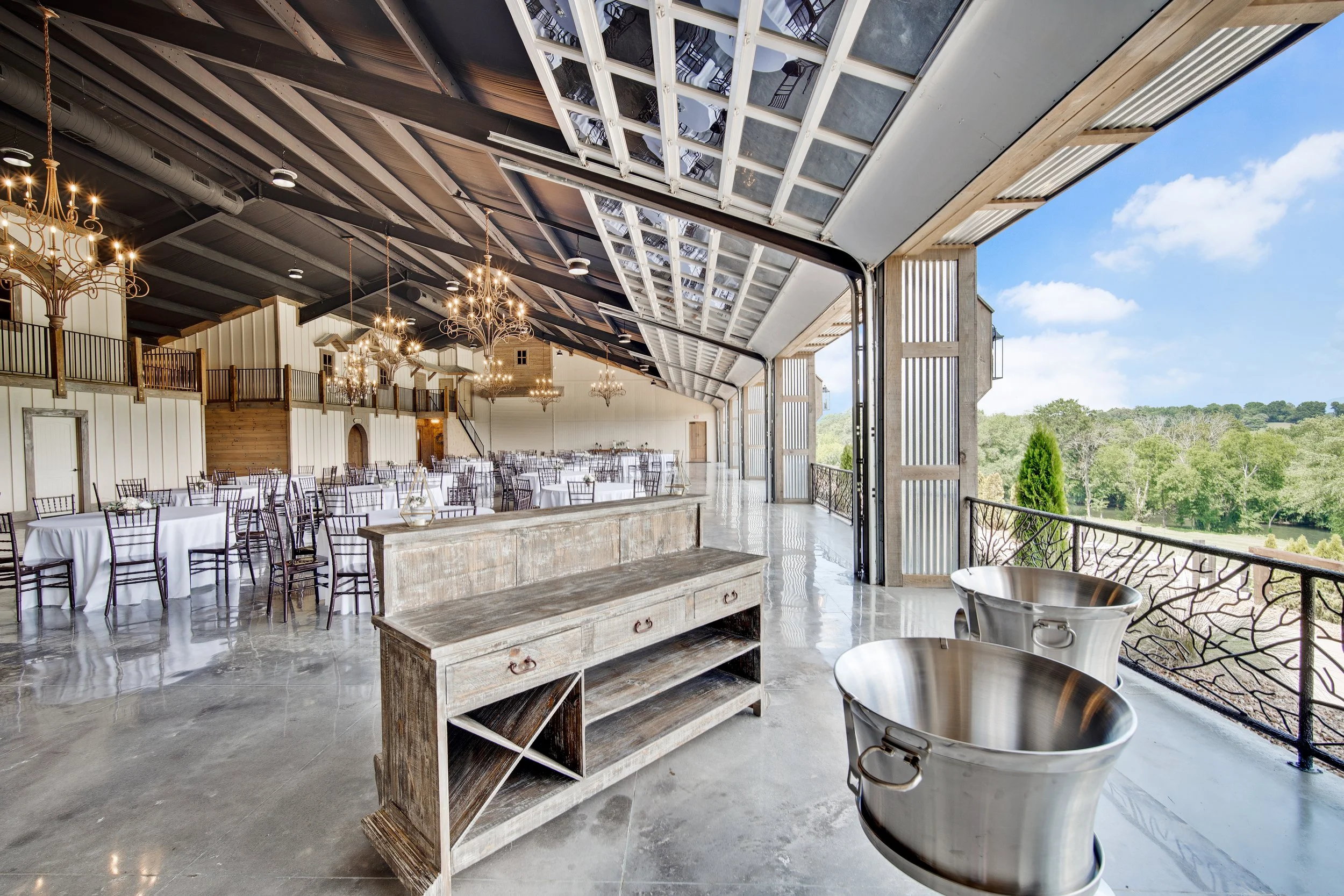 Open-air event space with round tables covered in white tablecloths, wooden chairs, hanging chandeliers, and a large open garage door showing a scenic view of green trees and blue sky.
