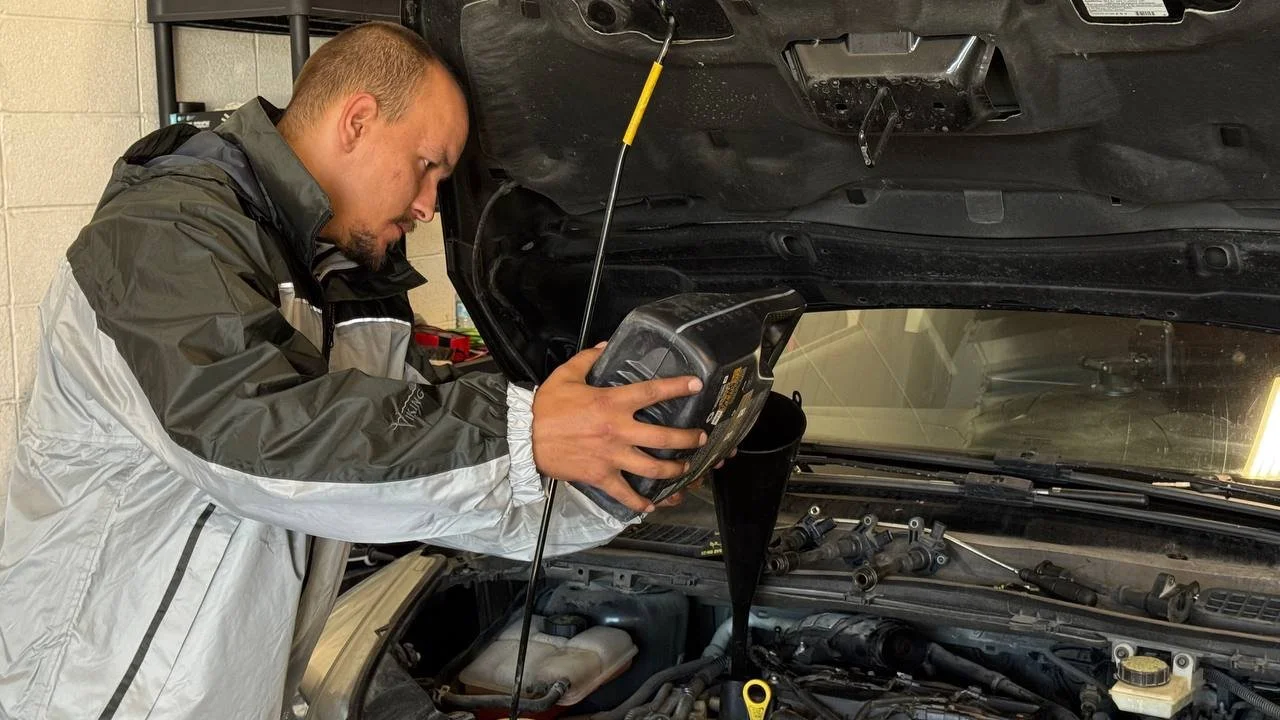 A man is refilling the oil in a car engine at an auto repair shop, pouring oil into the engine with the hood open.