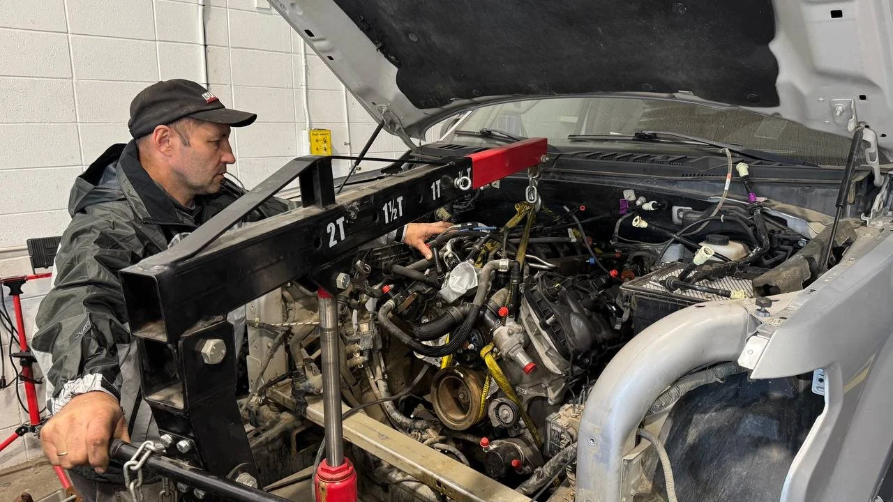 A man wearing a black jacket and cap inspecting a race car engine inside a garage, using a frame and measuring tools.