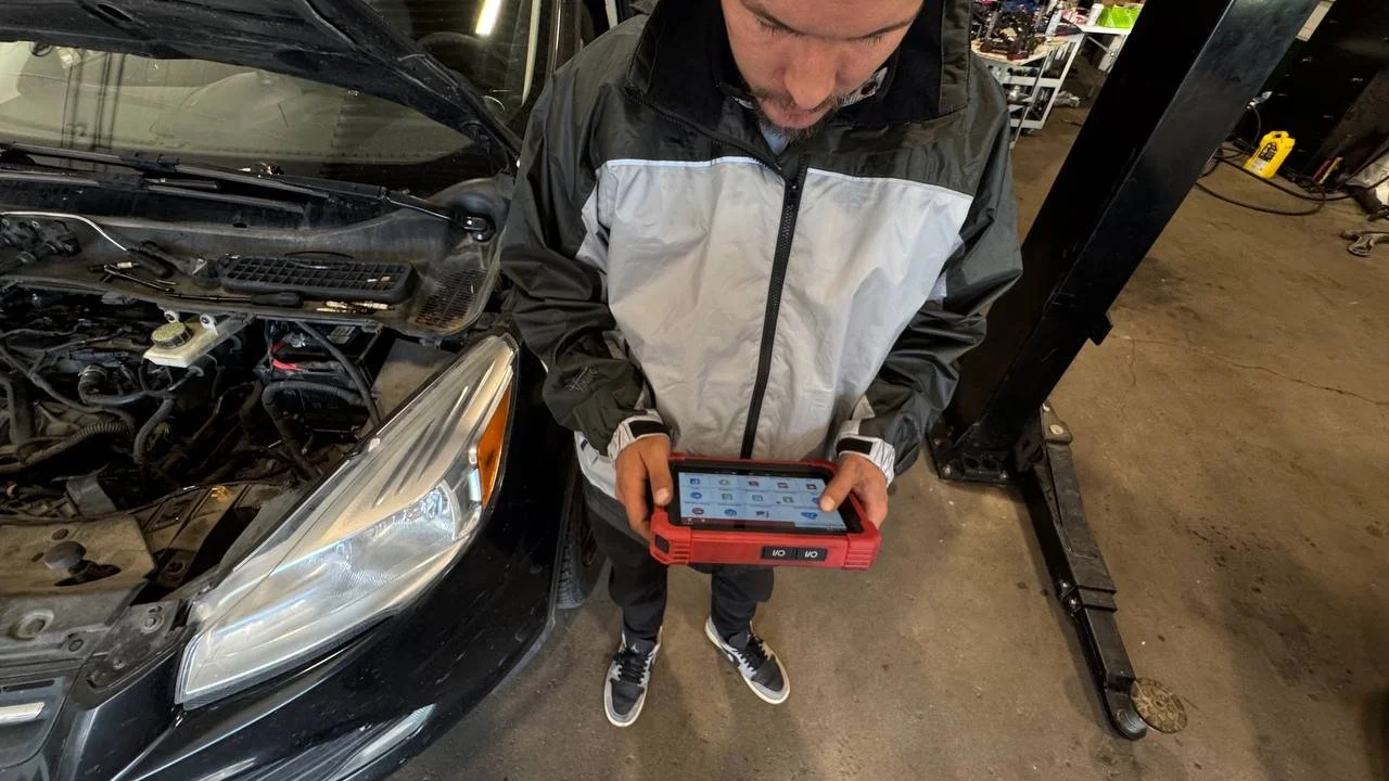 A man in a gray and black jacket stands beside a black car with its hood open, looking down at a diagnostic device in his hands at an auto repair shop.